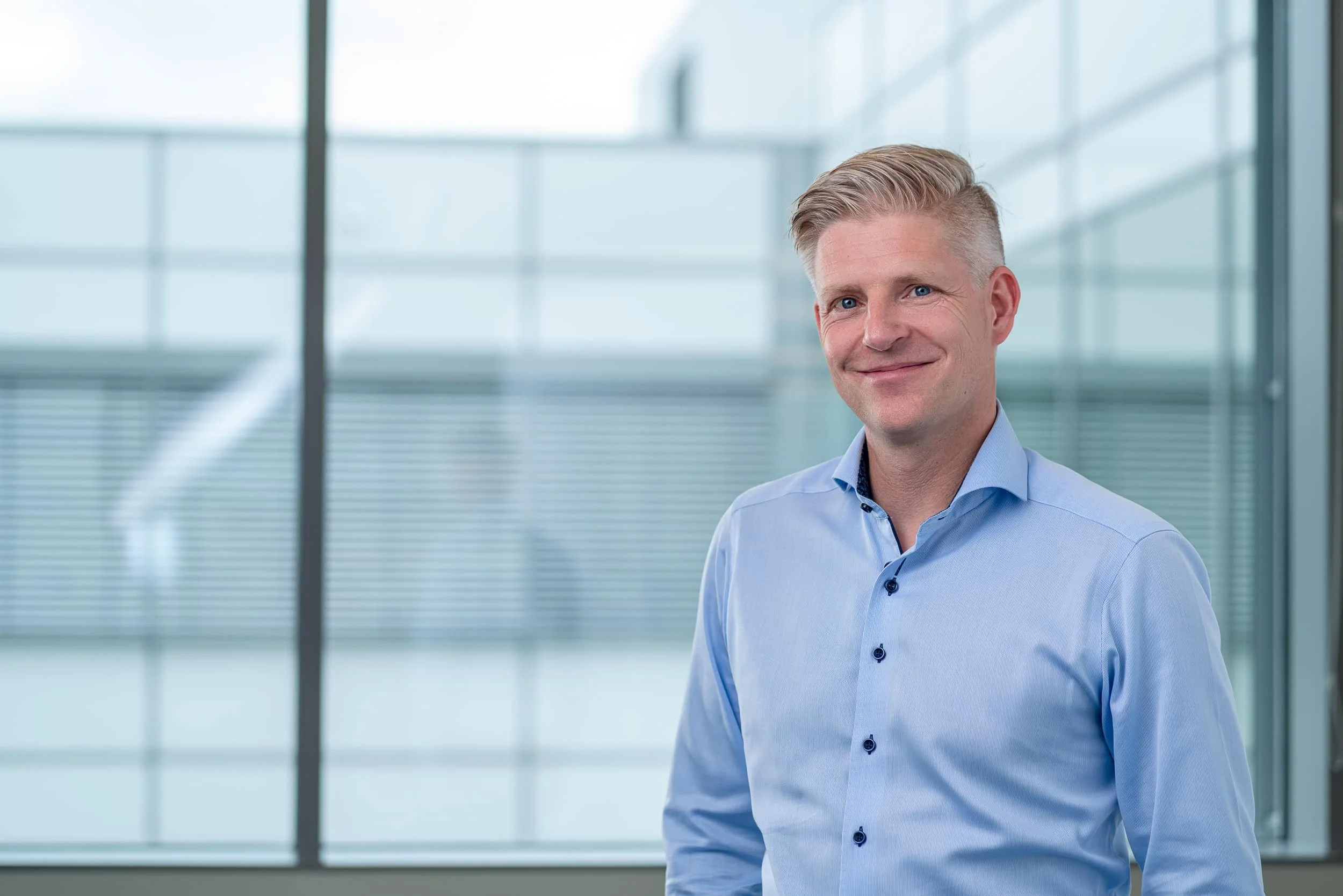 En mann med lyst hår og blå skjorte smiler og står foran et stort vindu i et moderne bygg.