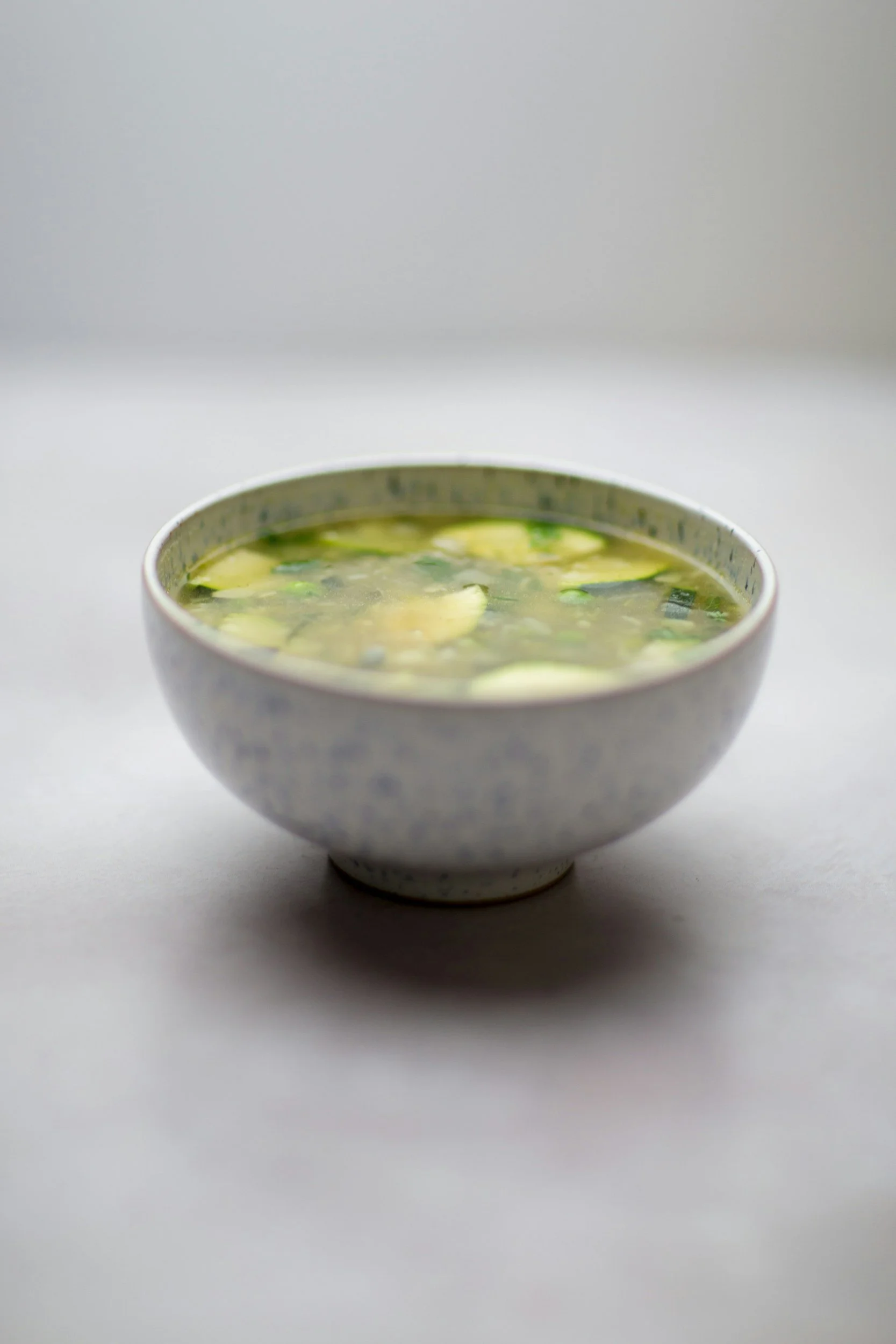 A ceramic bowl filled with Asian-style soup containing sliced vegetables and green onions, placed on a white surface.