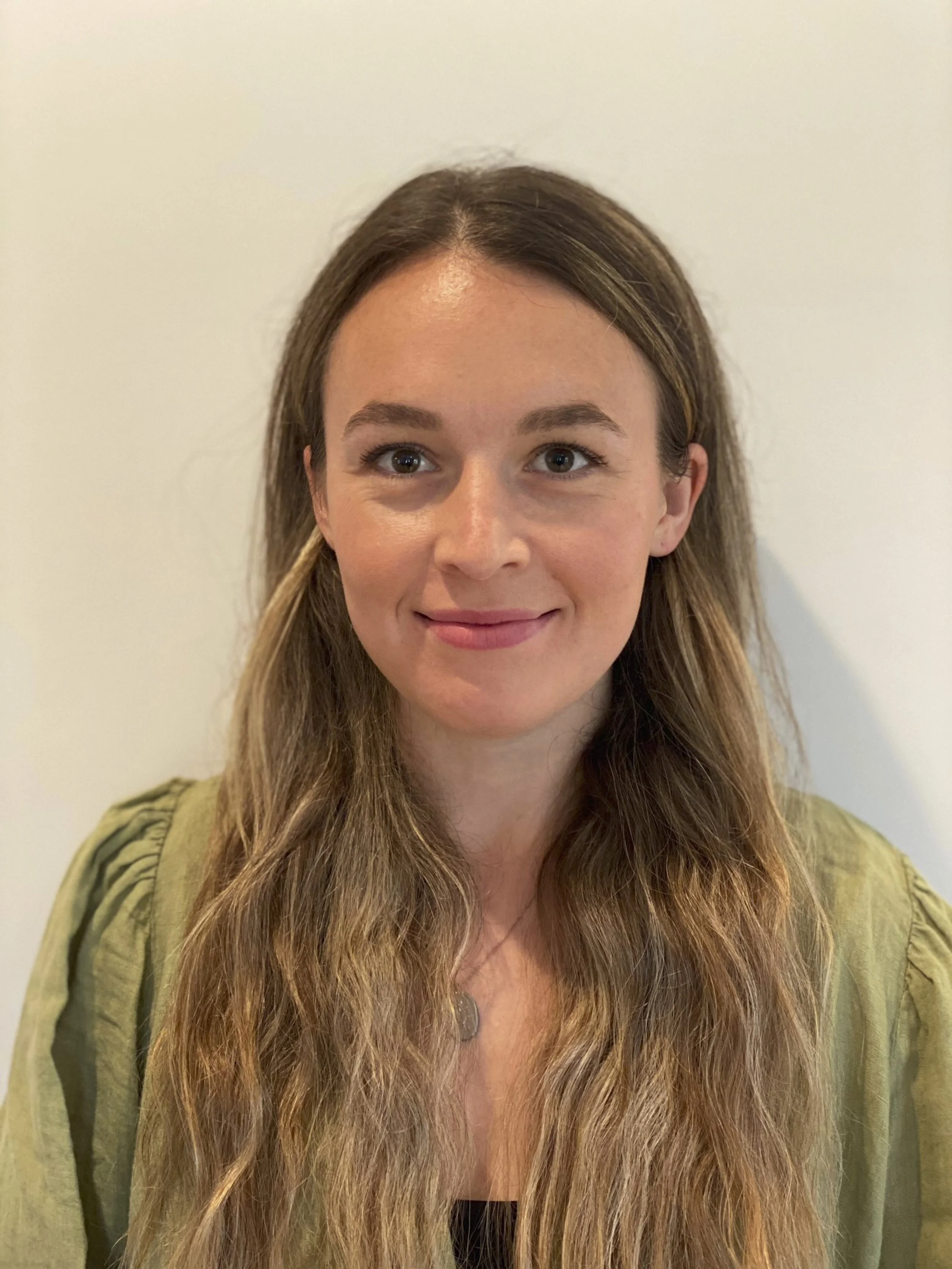 A woman with long, wavy brown hair and fair skin, smiling, wearing a green jacket and a necklace, standing against a plain light-colored background.