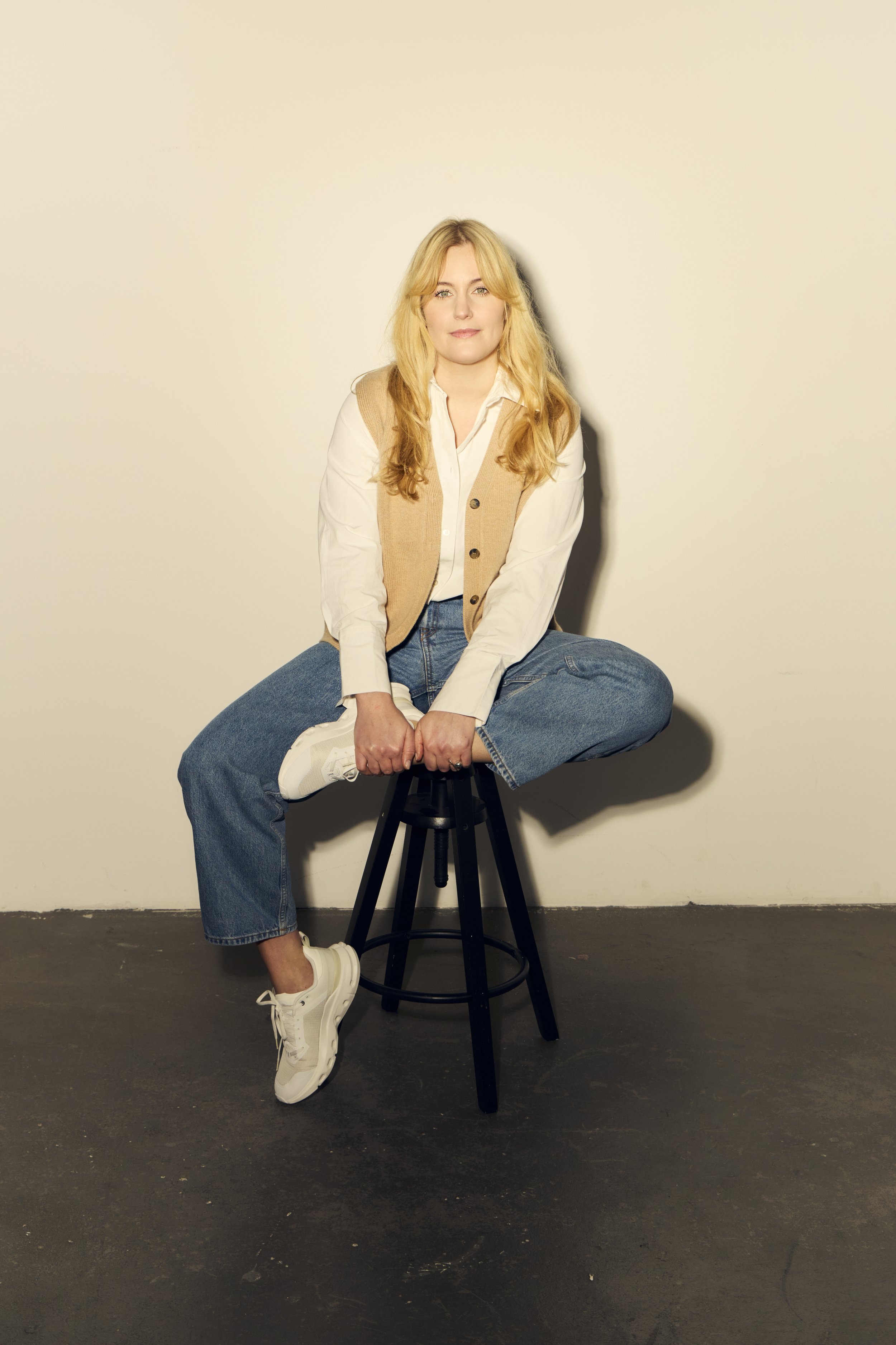 Weiße Frau sitzt auf einem Barhocker vor weißer Wand, trägt Jeans, weiße Schuhe, weißen Hemd und beigen Pullover und schaut selbstbewusst in die Kamera.