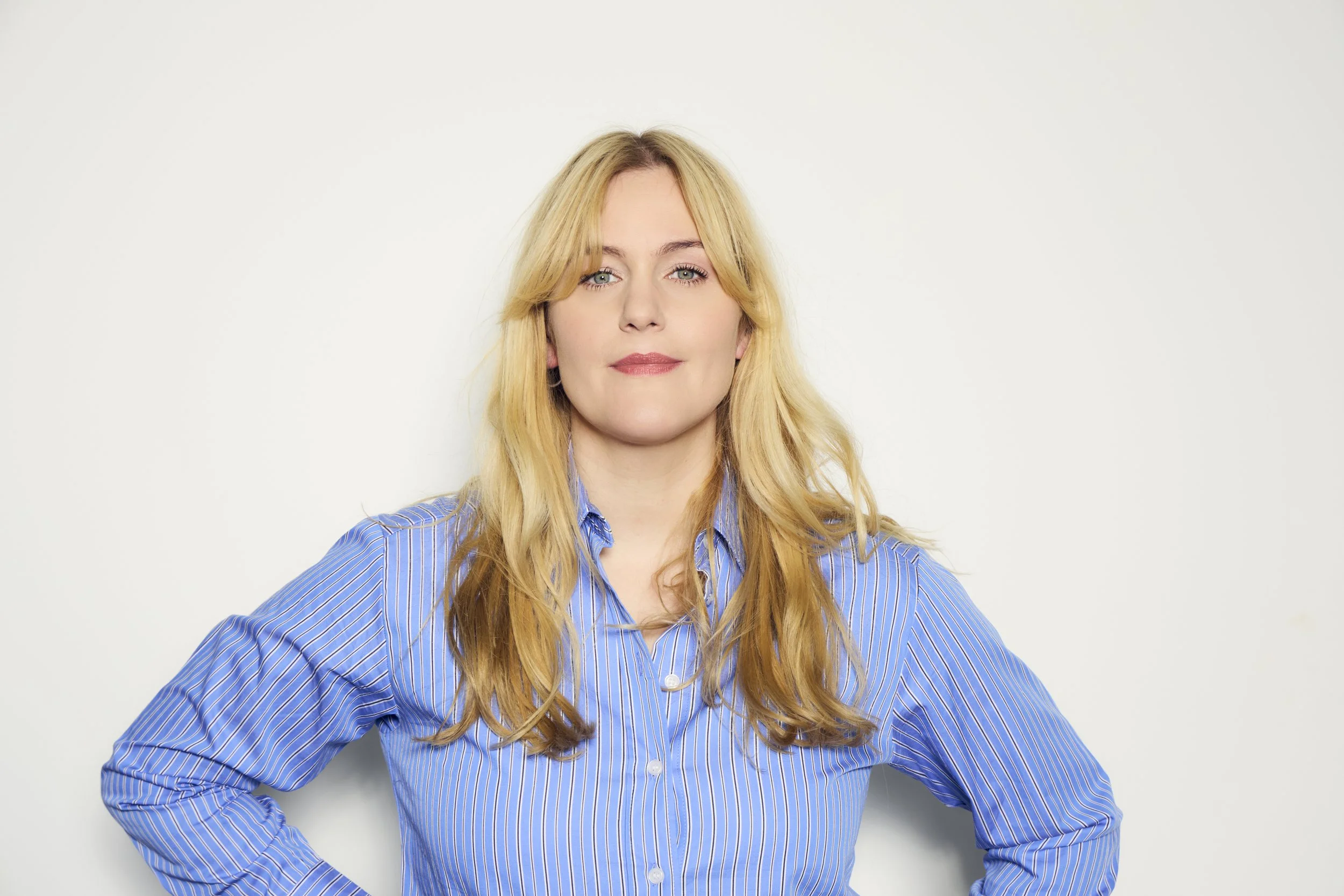 Weiße Frau trägt eine blaue, gestreifte Bluse, steht vor weißem Hintergrund und schaut empathisch, aber selbstbewusst in die Kamera.