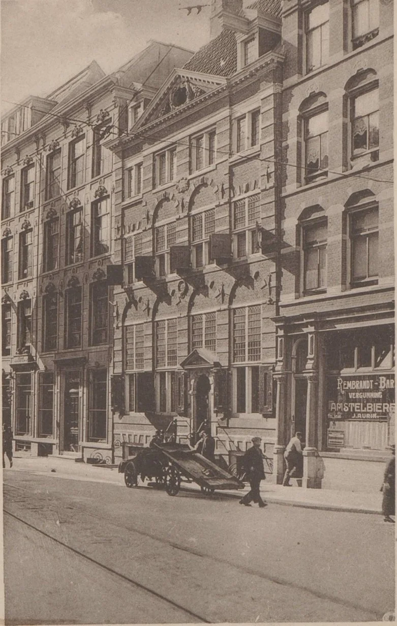 A black-and-white photograph of a vintage street scene in front of a multi-story building with ornate architecture, including arched windows and decorative cornices. Pedestrians are walking on the sidewalk, and there is a horse-drawn carriage in the center of the street.