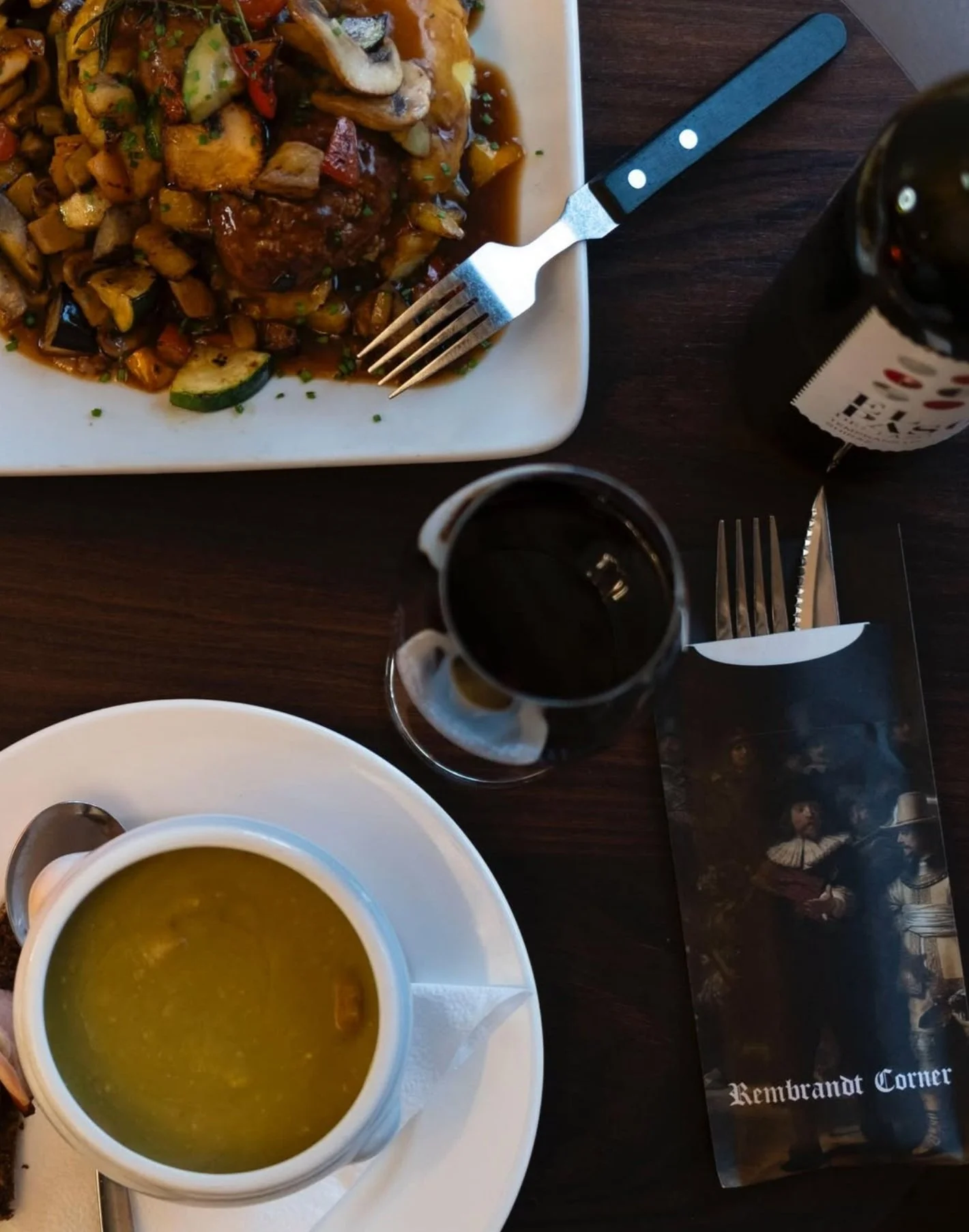 A meal setup with a bowl of green soup on a white plate, a cup, a glass of dark soda, a bottle of dark soda, and a plate of mixed vegetables with a fork, all on a dark wooden table.
