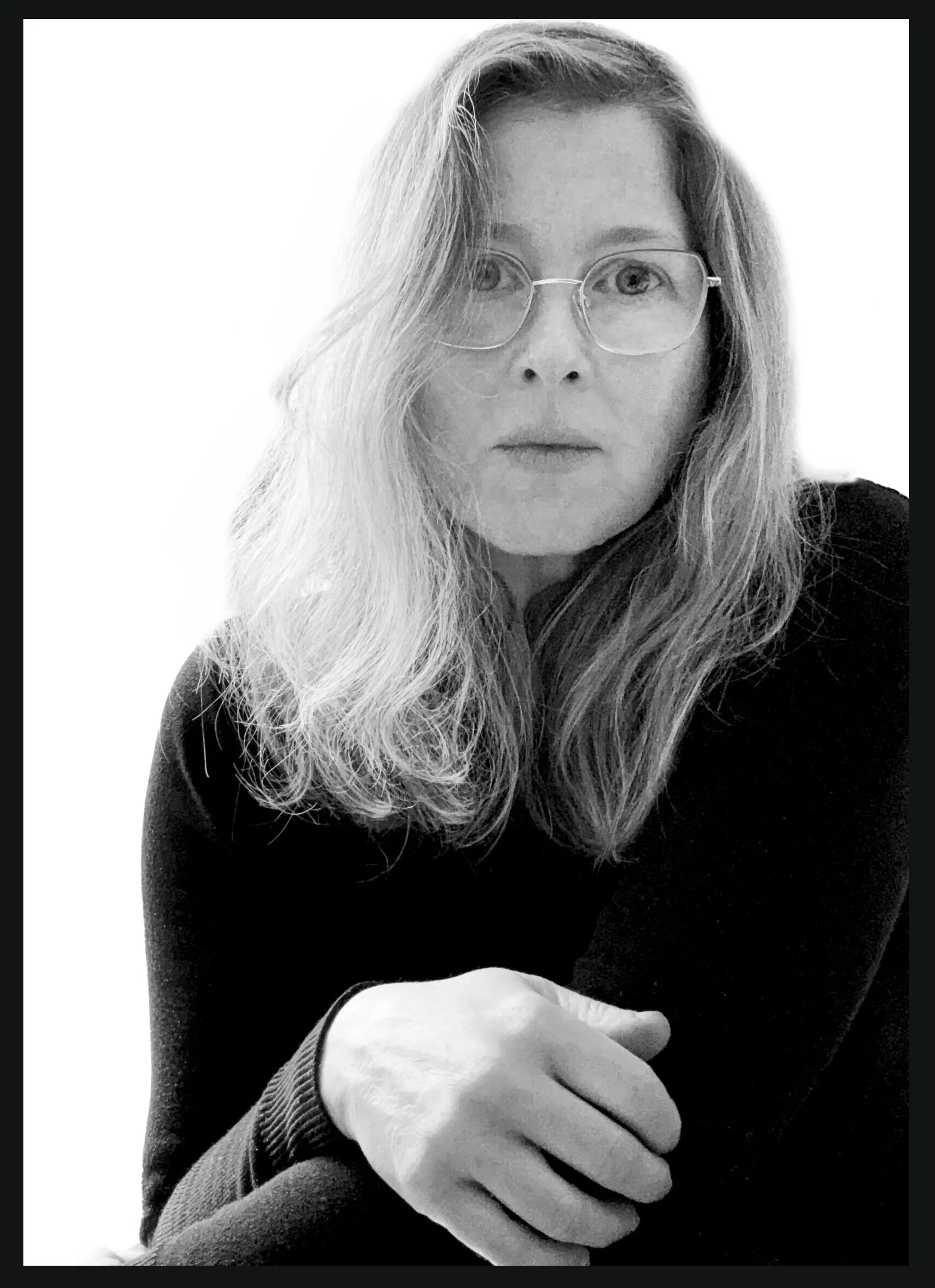 Black and white photo of a woman with long, wavy hair wearing glasses and a dark top, looking directly at the camera with a neutral expression.