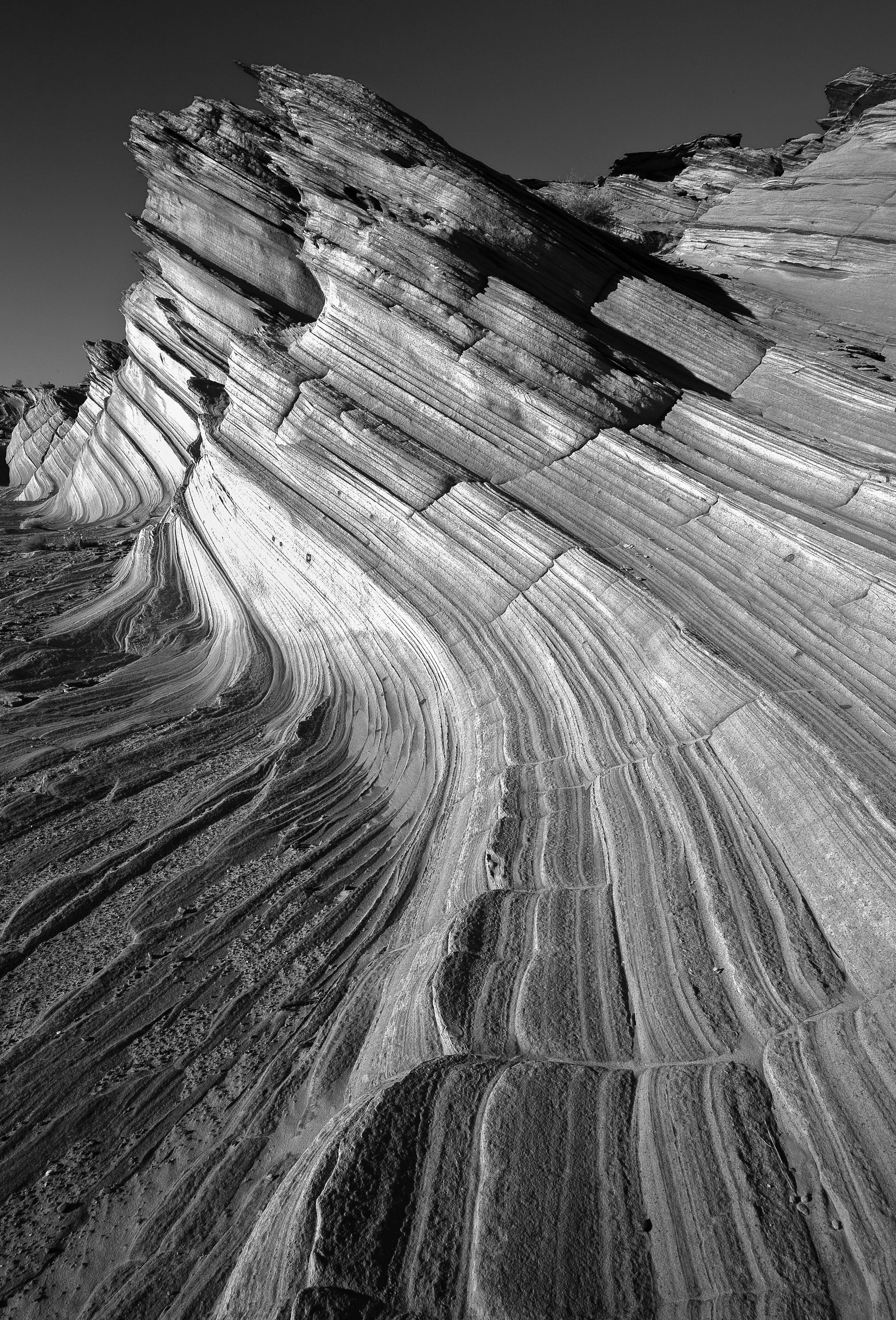 Black and white photograph of layered, striated rock formations with curved and jagged edges, likely in a desert or canyon landscape.