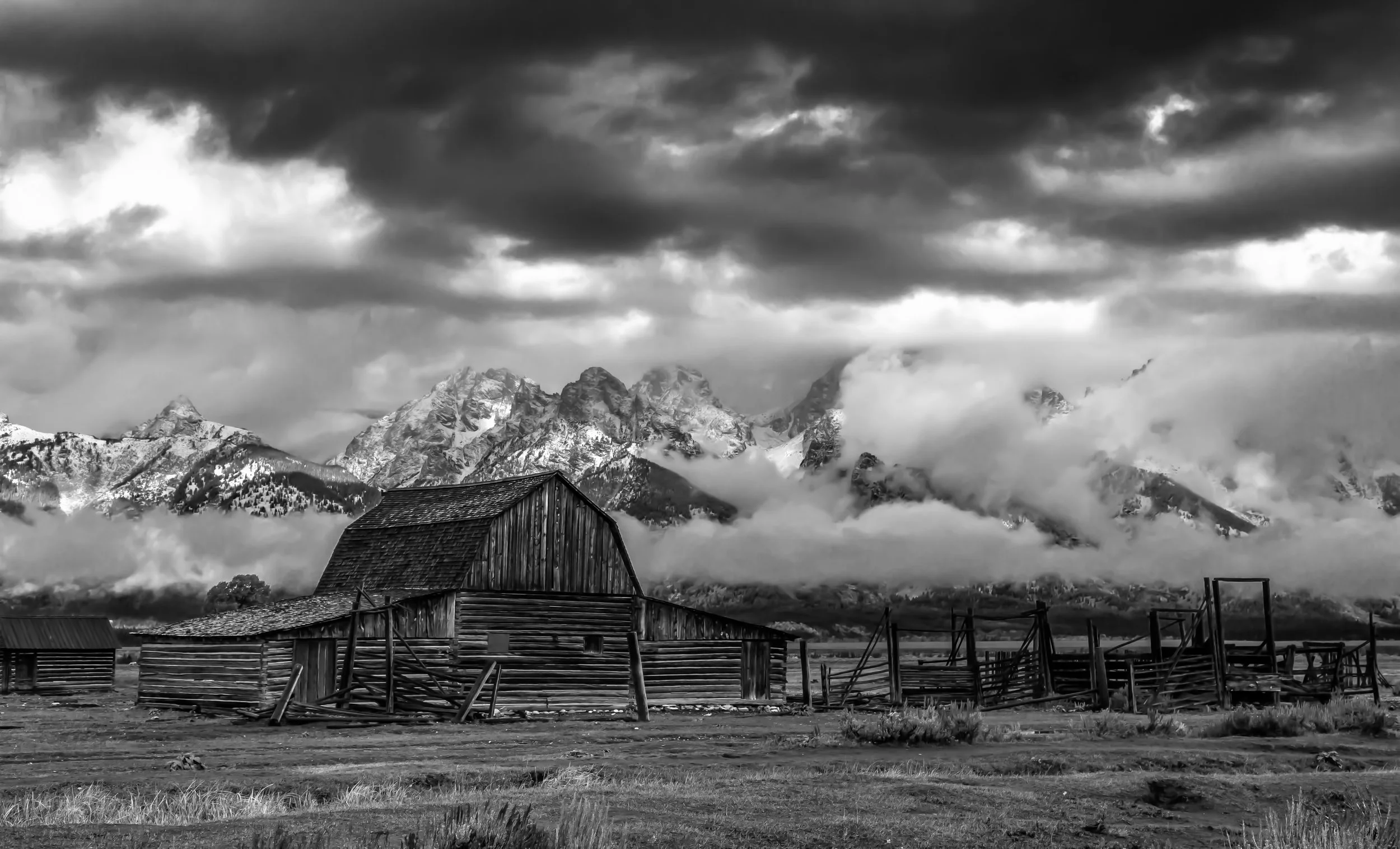 Black and white photo of a rustic barn in a rural landscape with mountain peaks and dramatic cloudy sky in the background.
