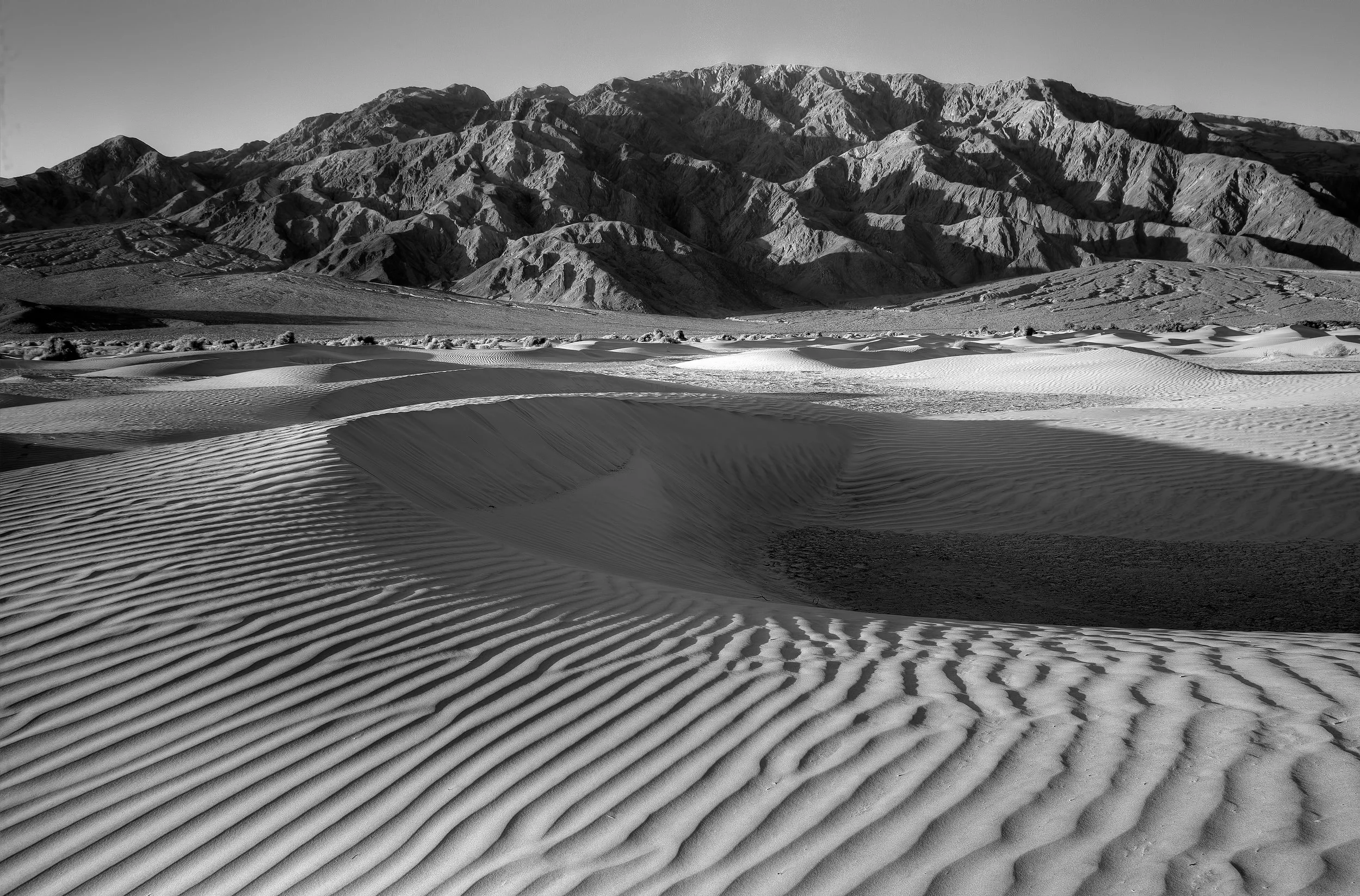 Black and white photograph of desert sand dunes with mountain range in the background.