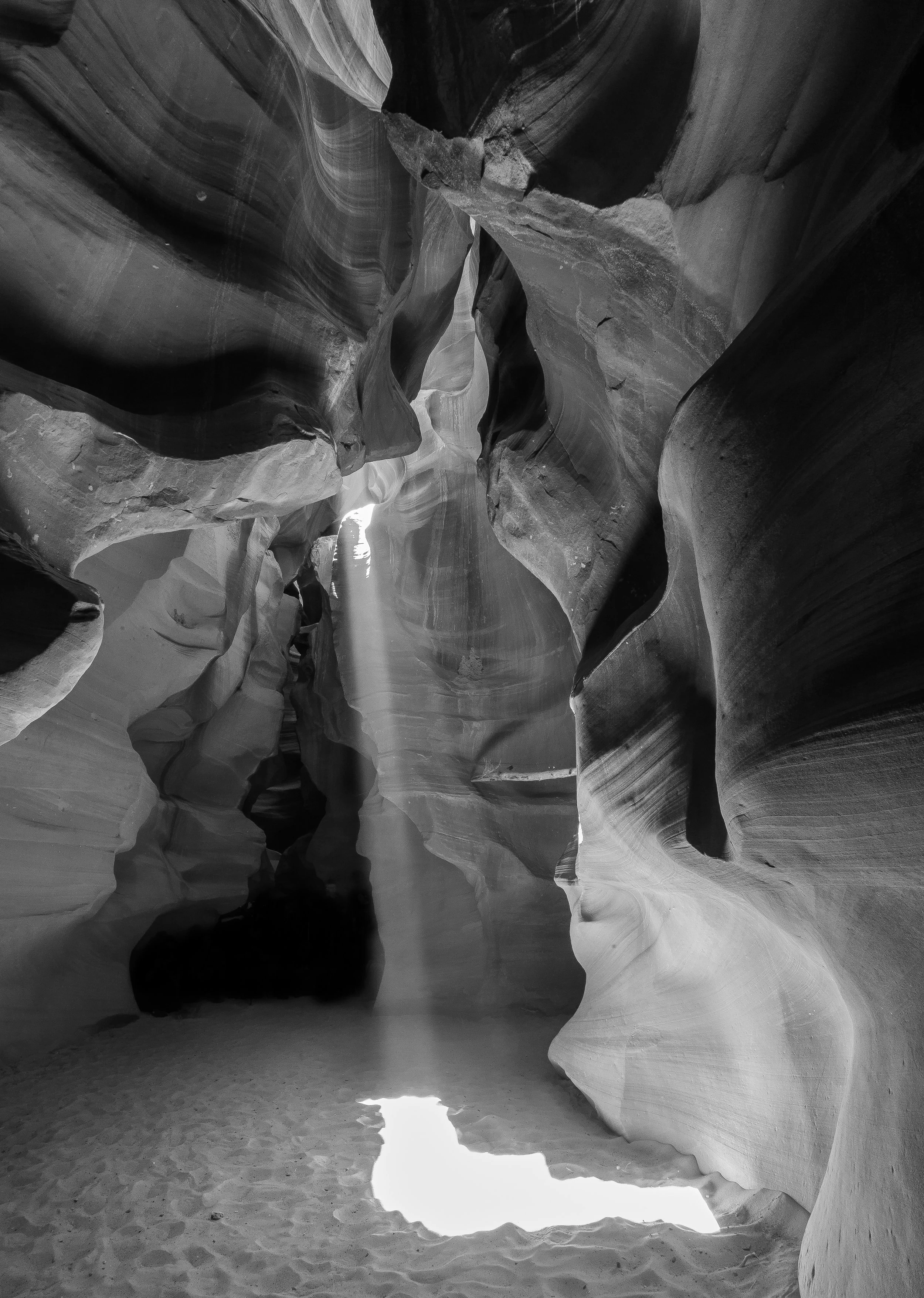 Black and white photo of a narrow slot canyon with smooth, wavy rock walls and sunlight shining through a crack at the top, illuminating the sandy floor.