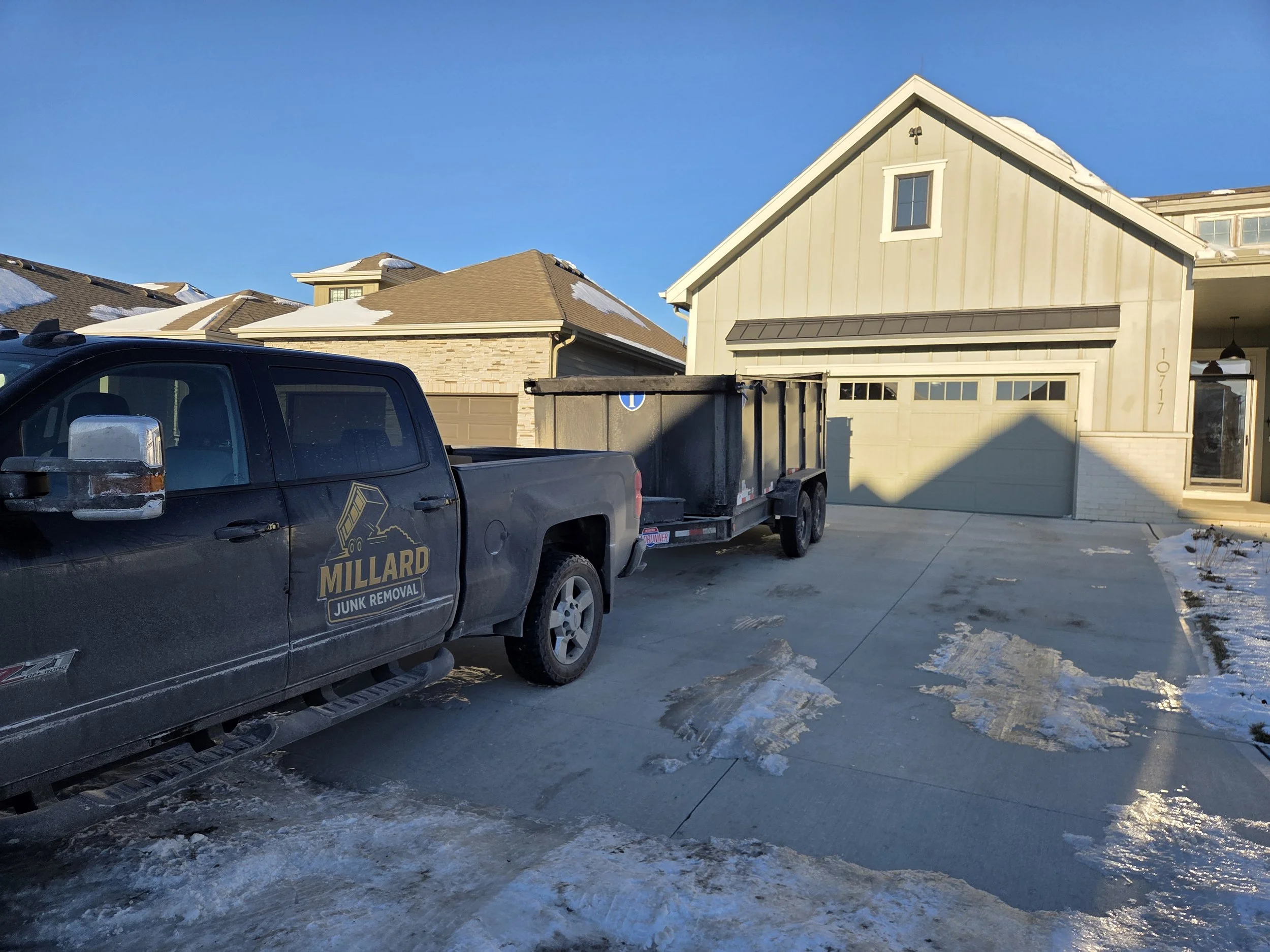 A Millard Junk Removal truck with a trailer parked in front of a light-colored house on a driveway with ice and snow, during the daytime with clear blue sky.