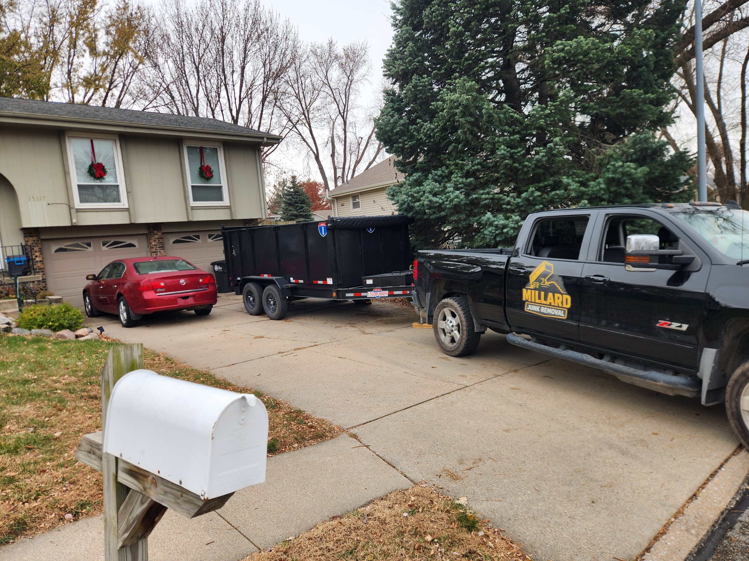 A residential driveway with a red car parked next to a house decorated with holiday wreaths, a black Millard Junk Removal truck and trailer backing up, and a large evergreen tree in the background.