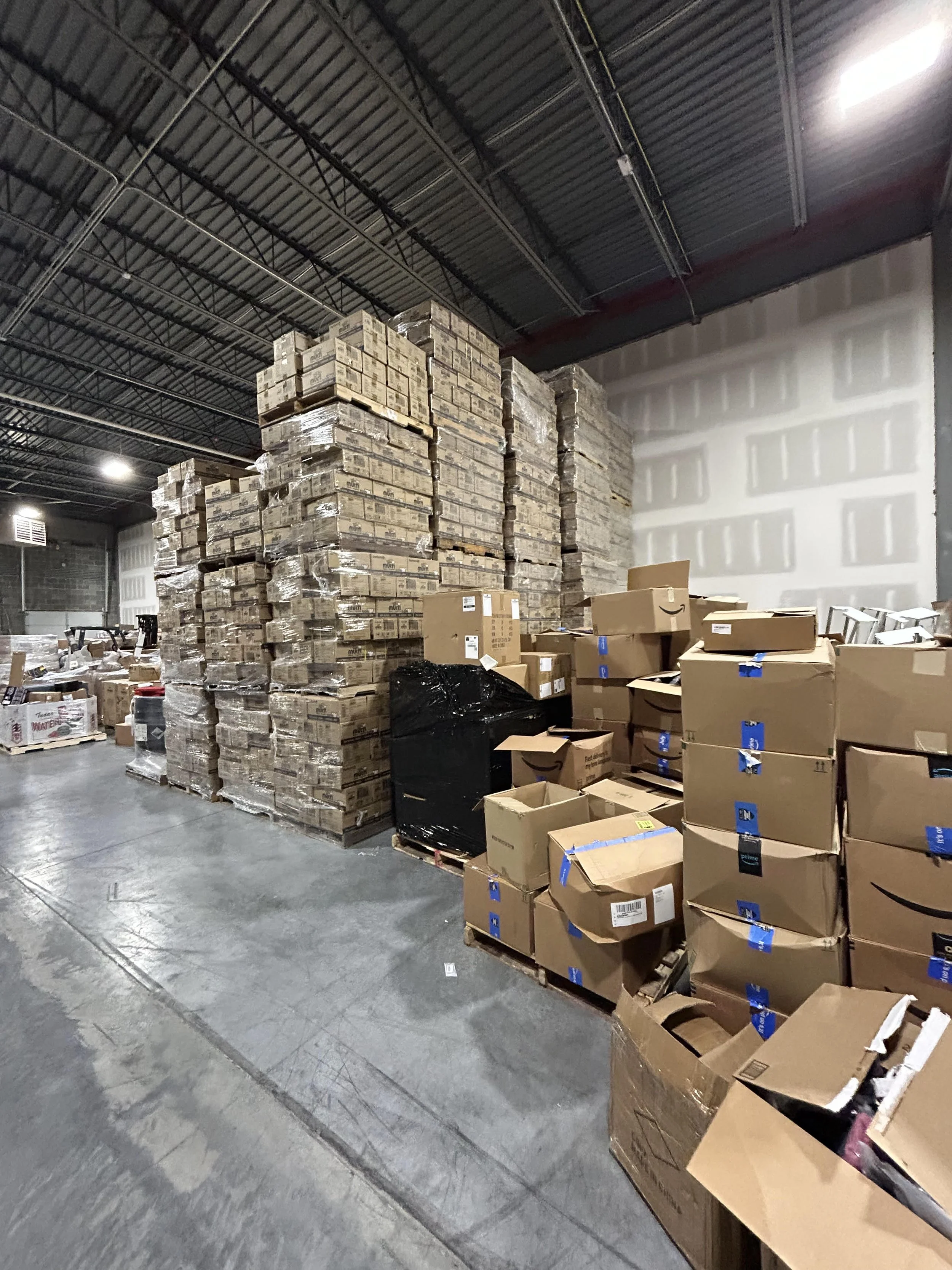 Interior of a warehouse with stacks of cardboard boxes and pallets, some wrapped in plastic, on the concrete floor before a Millard Junk Removal commercial cleanout job.