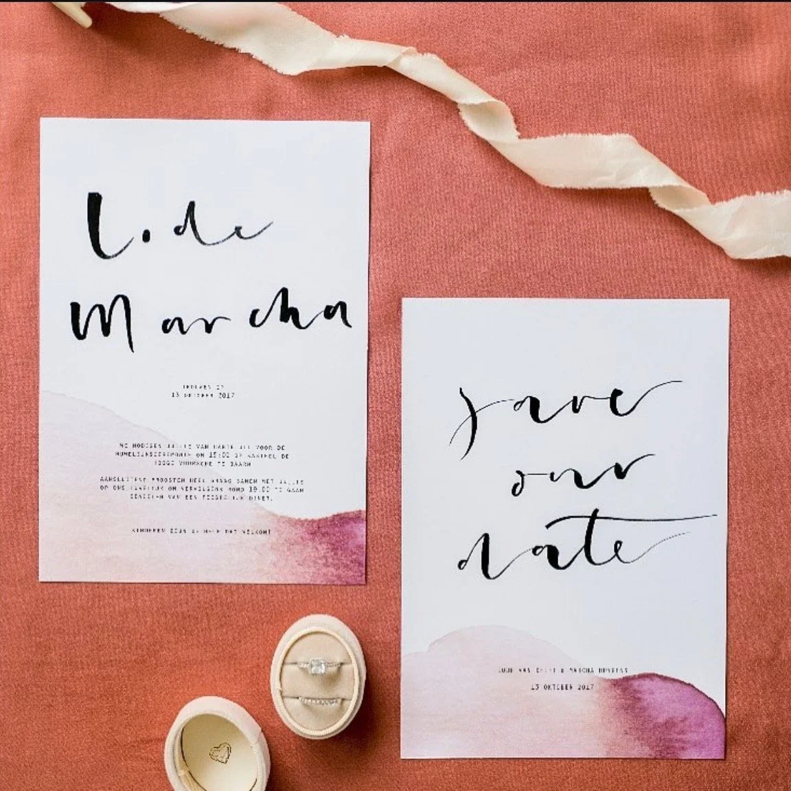 Save the date kaartjes met roze en lavendelkleurige waterverf achtergrond, handgeschreven zwart lettertype, inclusief een ringdoos met twee ringen en een wit fotolijstje, allemaal op een roestkleurige stoffen achtergrond.