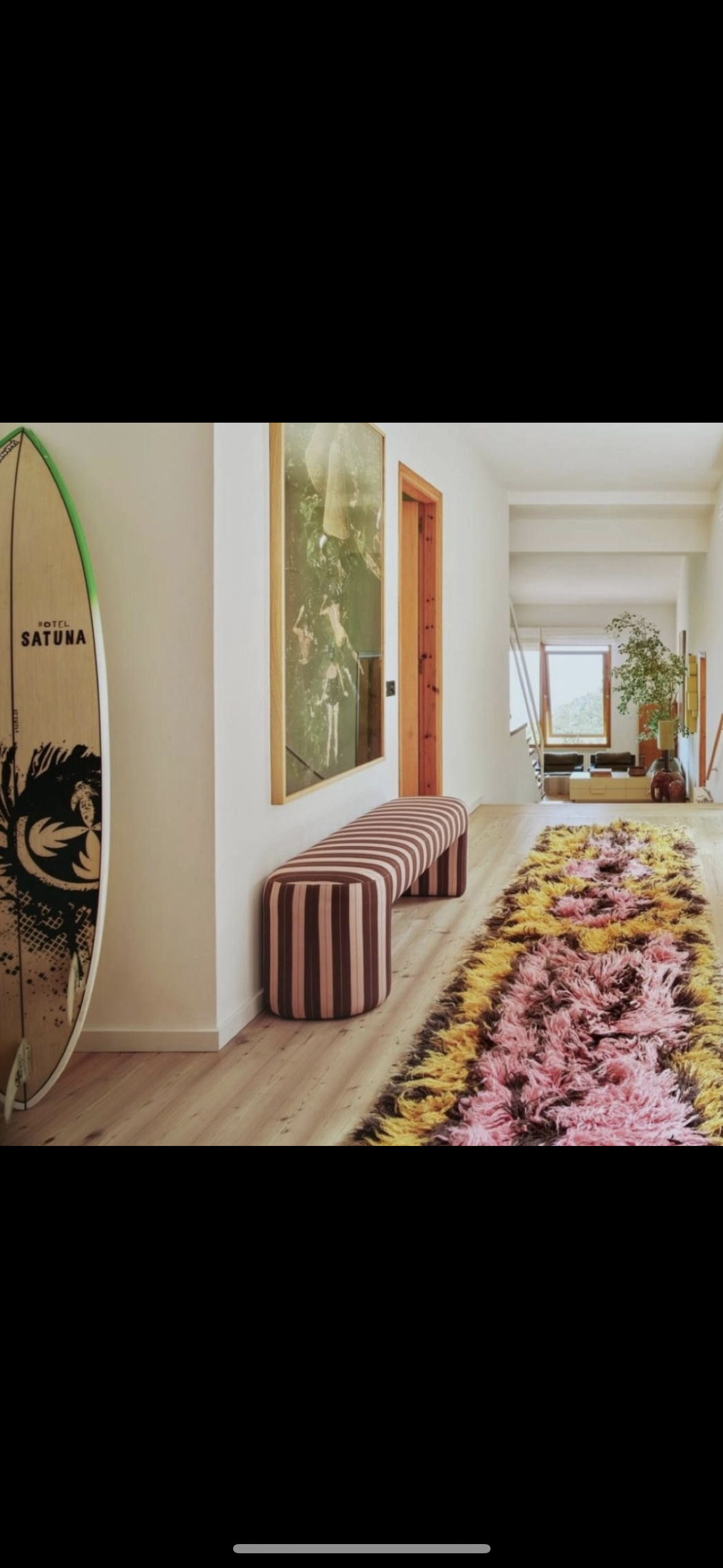 Interieur van een woning met een smalle, kleurrijke tapijtrug, een gestreepte bank, een surfplank, een kunstwerk en groene planten.