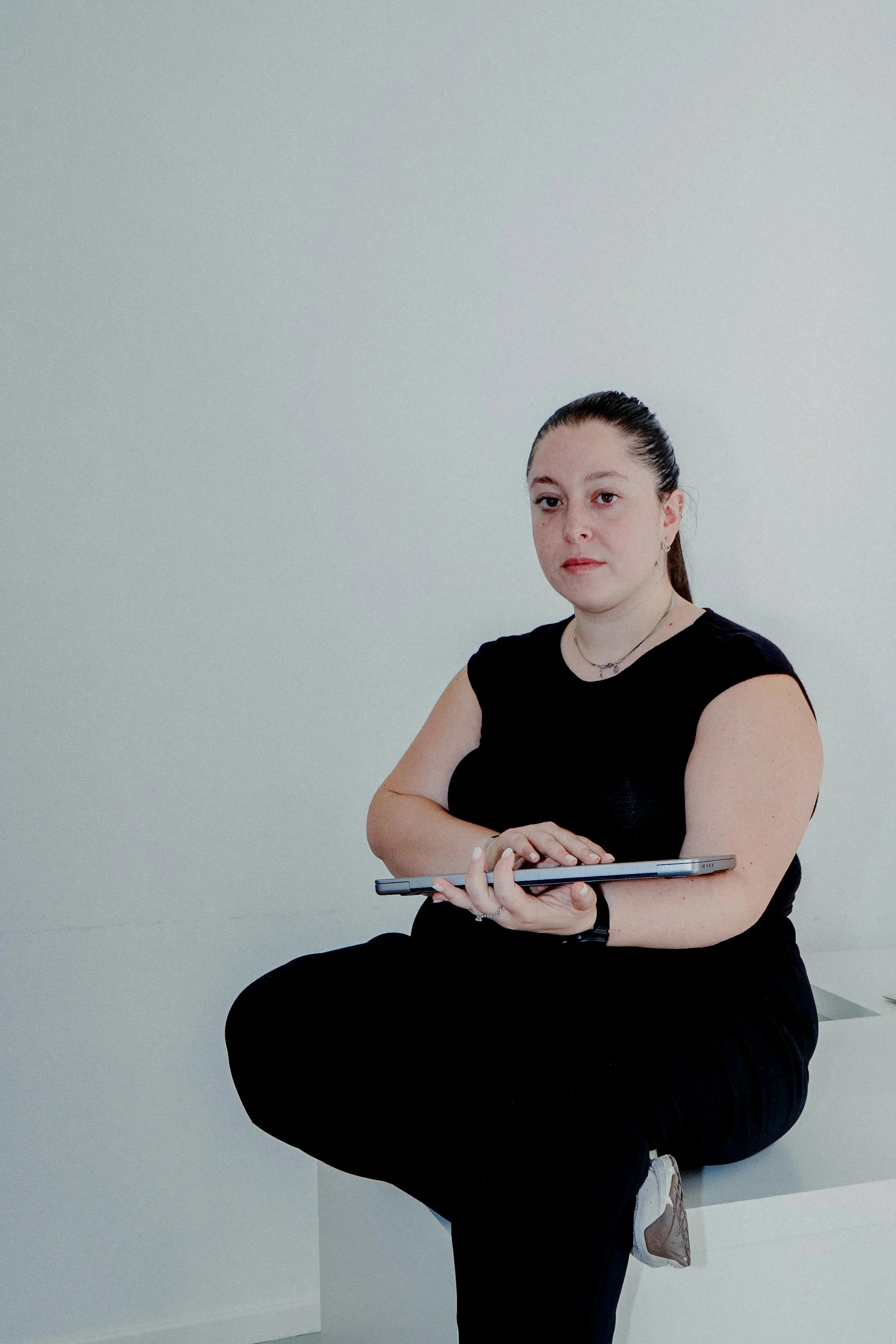 A man with dark curly hair, glasses, and a beard, smiling slightly. He is wearing a dark green sweater and a white shirt underneath. Behind him is a blurred background with a plant visible on the right side.