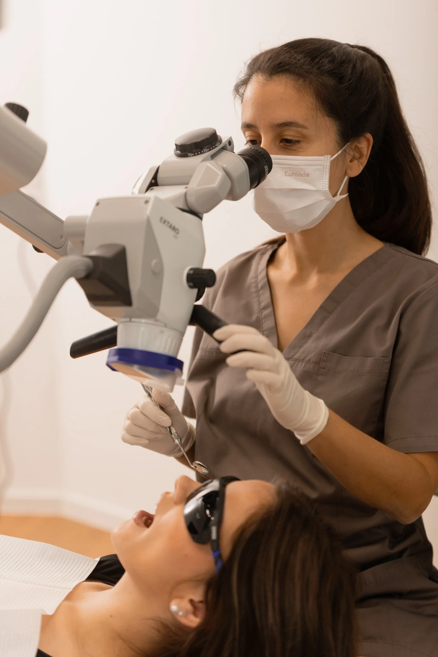 Zahnärztin Dr. Sina Klai mit Schutzmaske und Handschuhen untersucht eine Patientin mit einer Lupe unter einem Mikroskop.