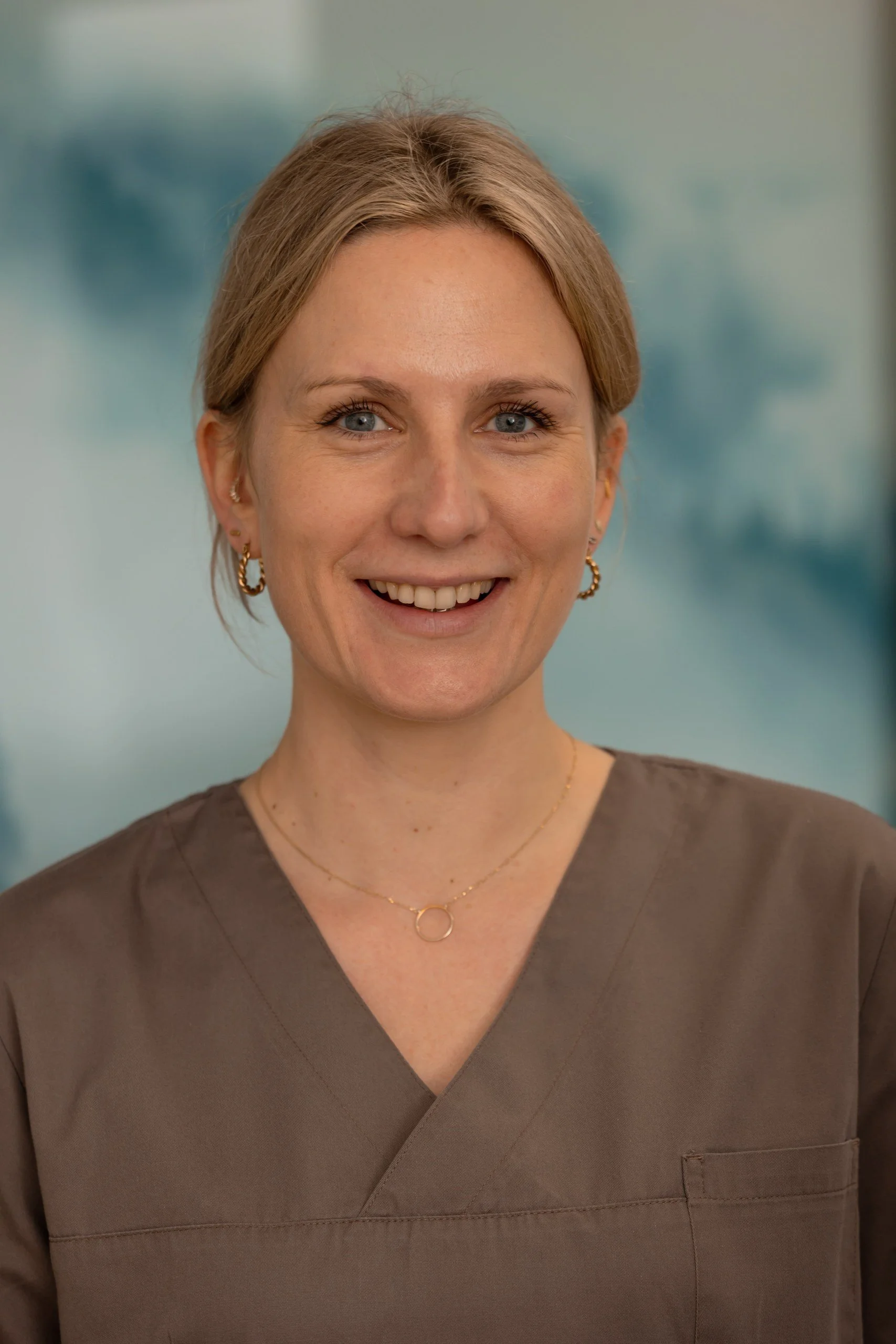 Carla Böhm unsere Zahnärztin für Vorsorge trägt braune Arbeitskleidung und trägt Goldschmuck, sie lächelt vor einem unscharfen blauen Hintergrund.