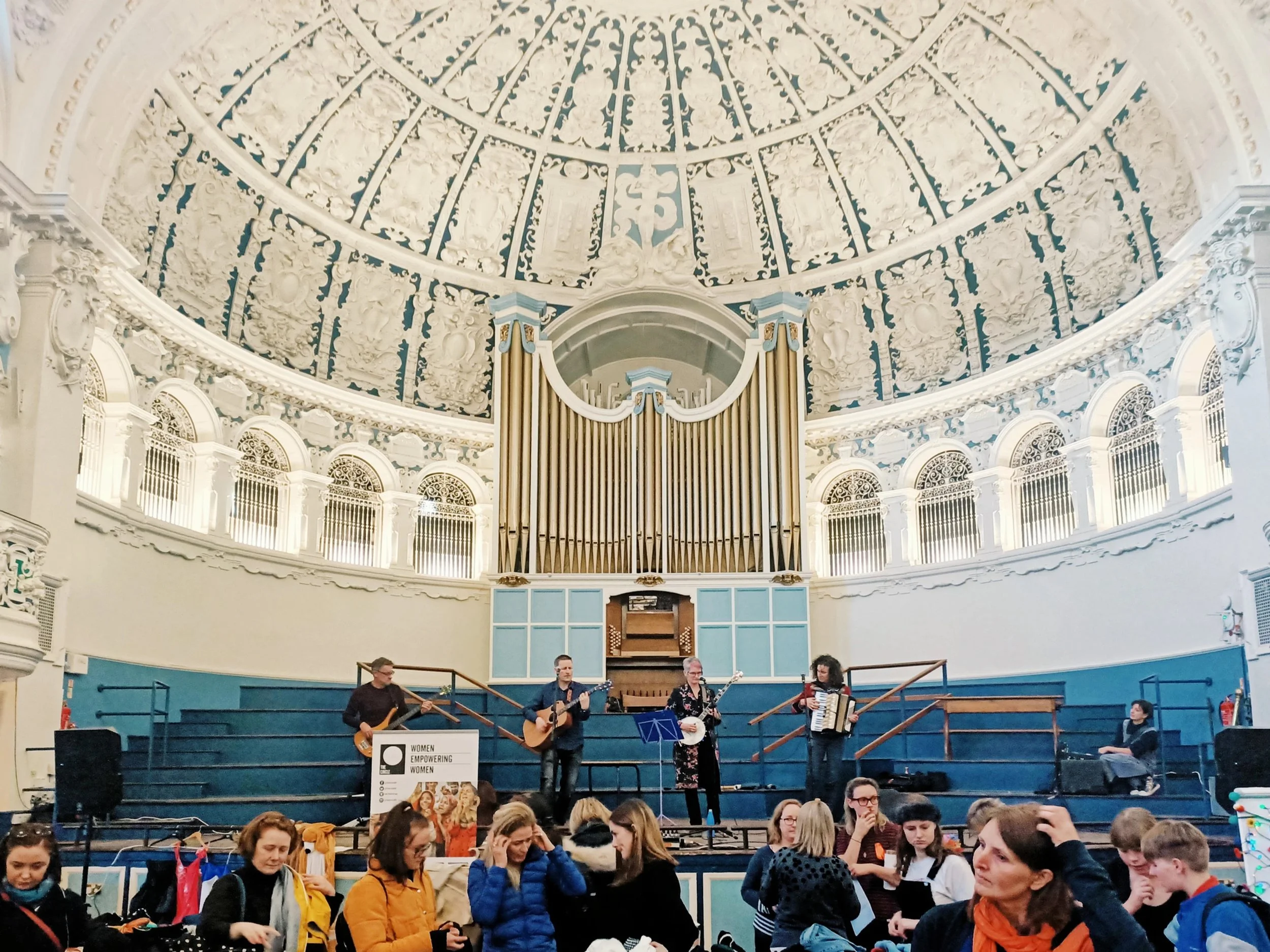 A band performing on stage inside a grand, ornate hall with a high, decorated ceiling. The band consists of five members, playing guitars, drums, and an accordion, with an audience watching.