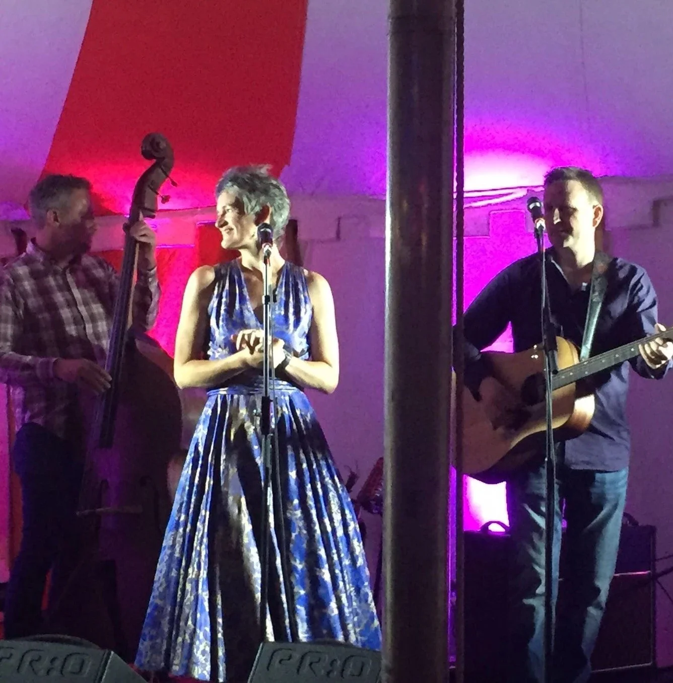 A musical performance with three performers on stage, a woman in a patterned blue dress in the center, a man with a guitar on the right, and another man with a double bass on the left, with colorful stage lighting.
