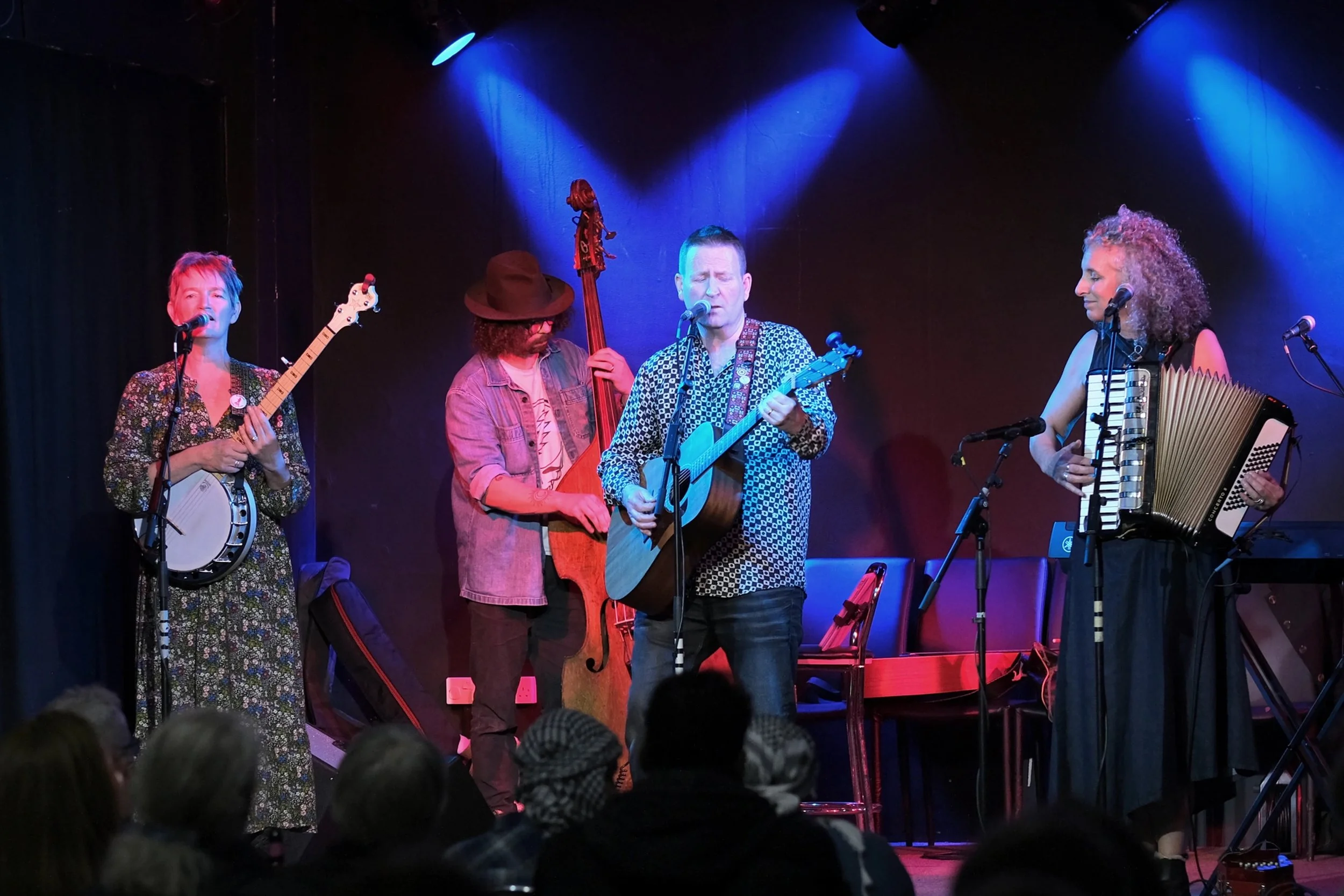 Four musicians performing on stage with various instruments, including a banjo, upright bass, guitar, and accordion, under blue and purple stage lighting.