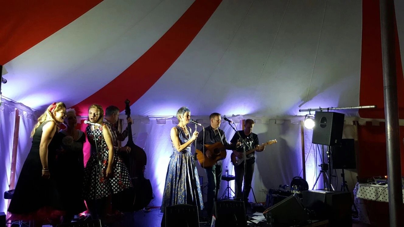 Musicians performing on stage in a tent, with three women singing and three men playing instruments, including acoustic and electric guitars, under colorful lighting.