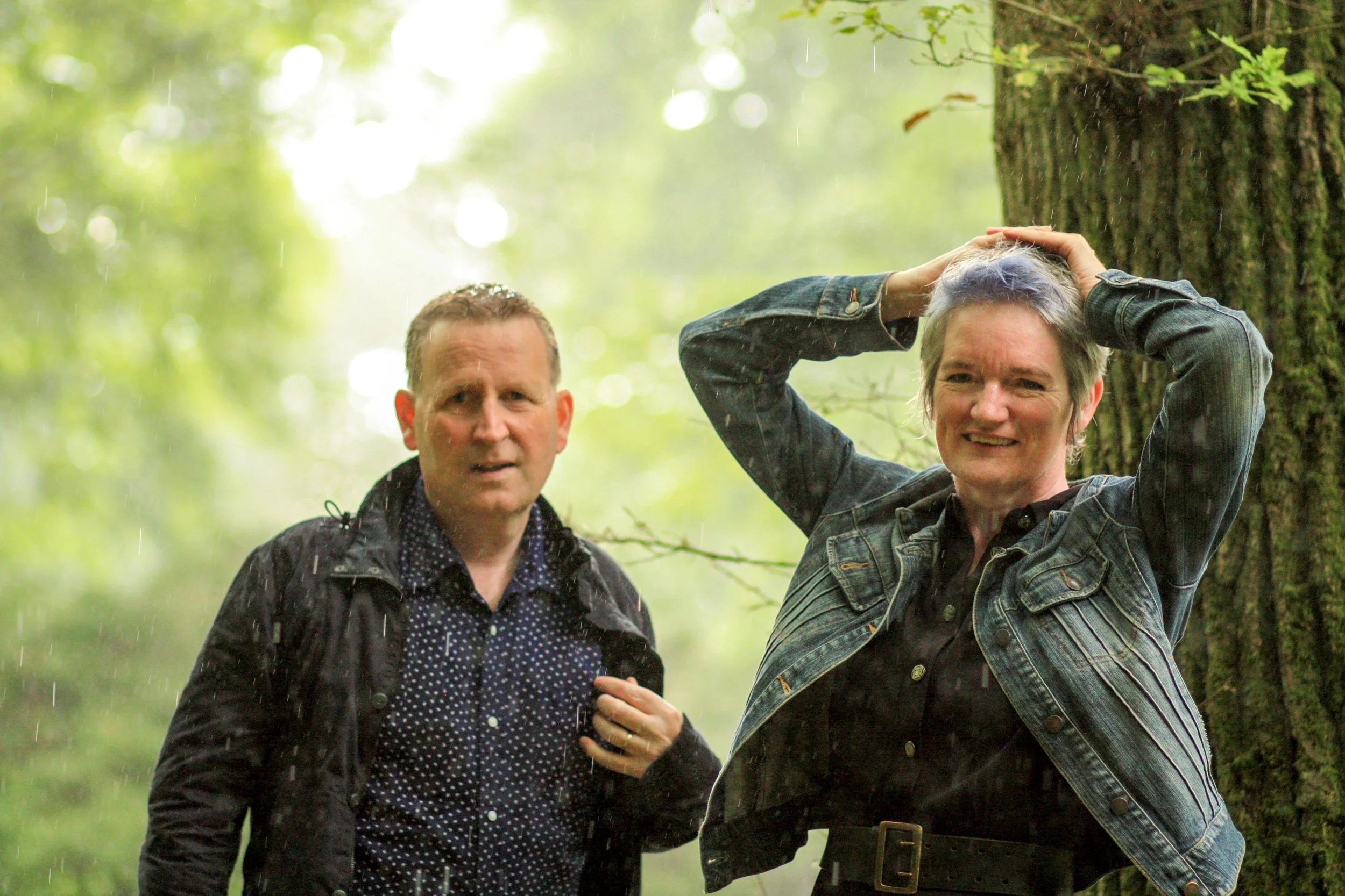 Two people standing outdoors next to a tree in the rain, one man and one woman, both appearing surprised or confused.