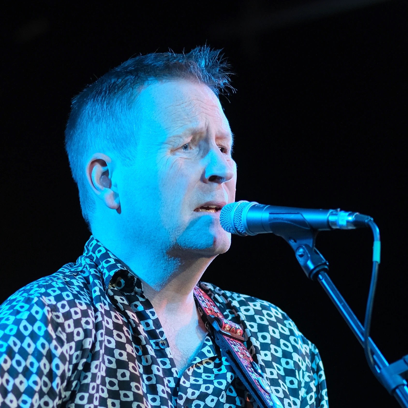 A man singing into a microphone on stage, wearing a patterned shirt with a guitar strap.
