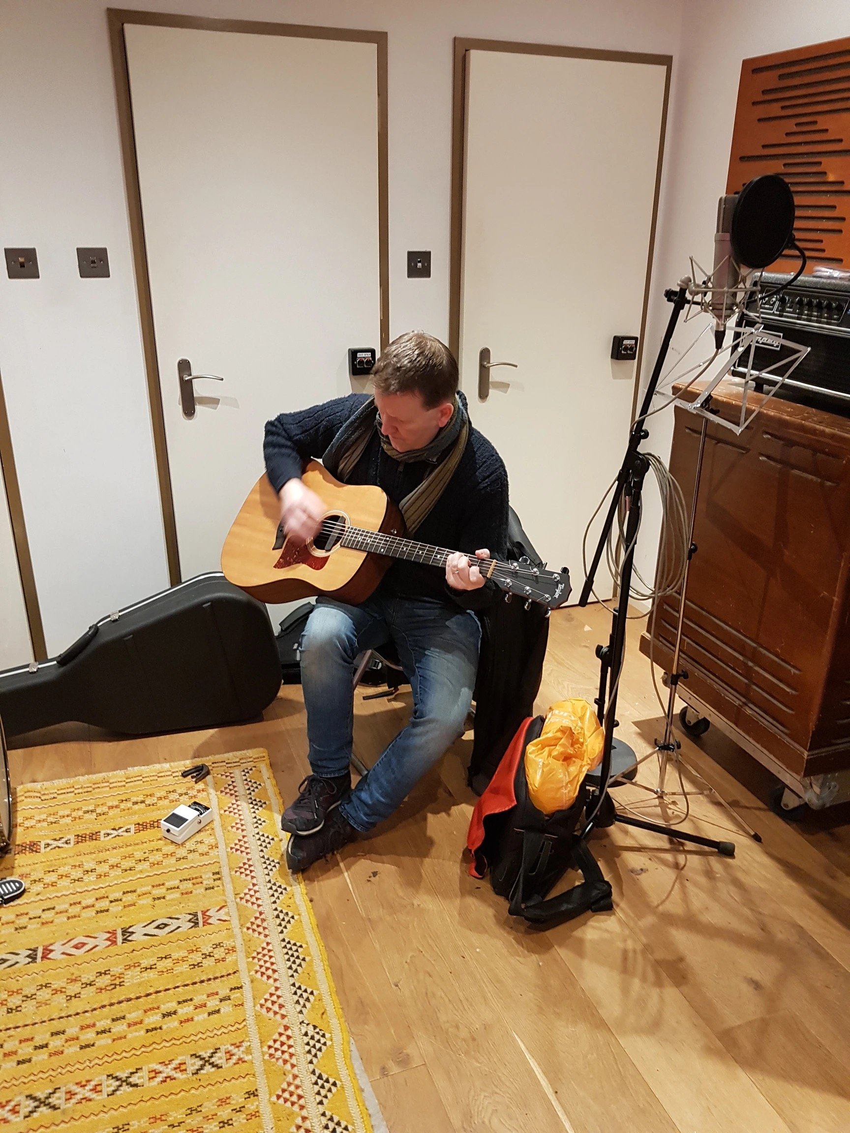 A man playing an acoustic guitar in a recording studio with wooden floors, a yellow patterned rug, and music equipment including a microphone on a stand and an amp. There are three white doors in the background.