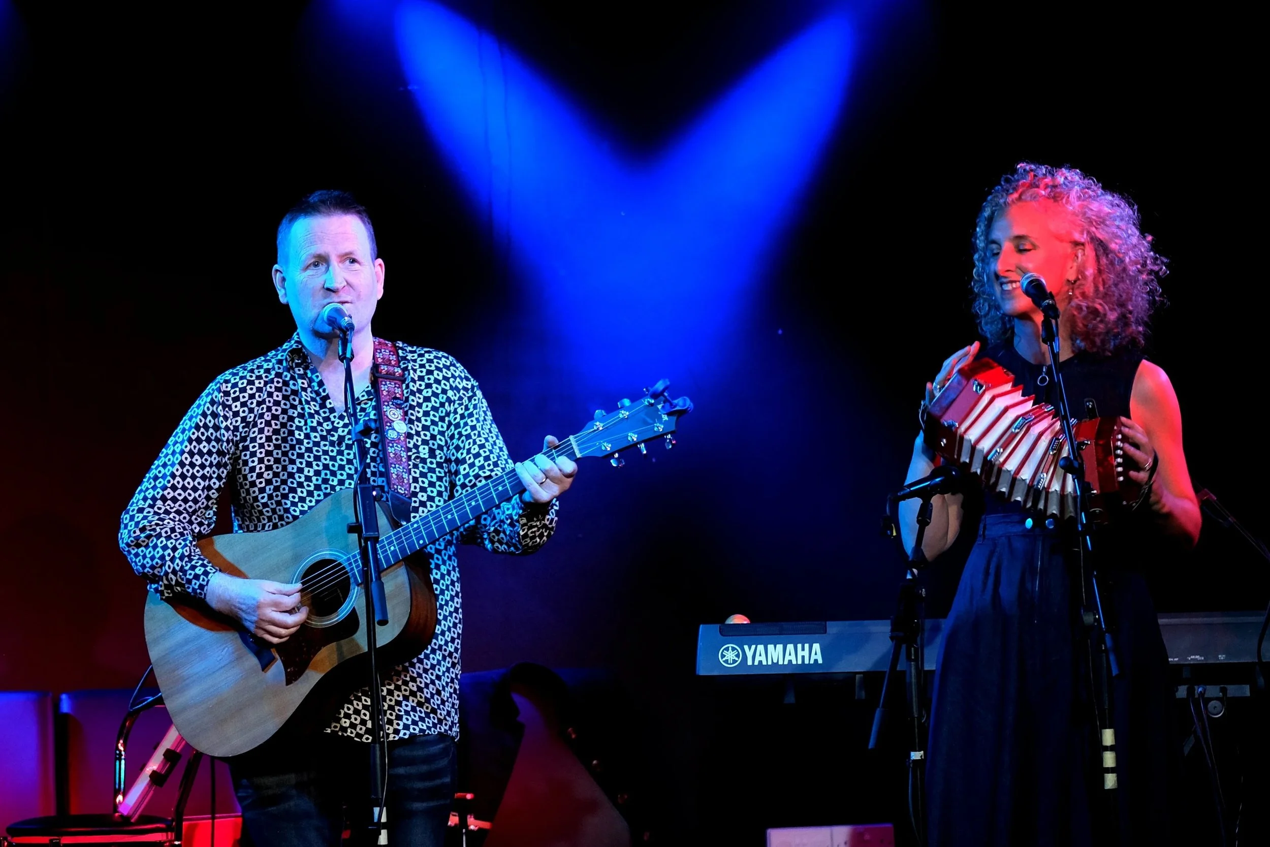 A man playing an acoustic guitar and singing into a microphone, and a woman singing into a microphone while holding a row of small melodicas on her chest, performing on a stage illuminated with blue and purple lighting.