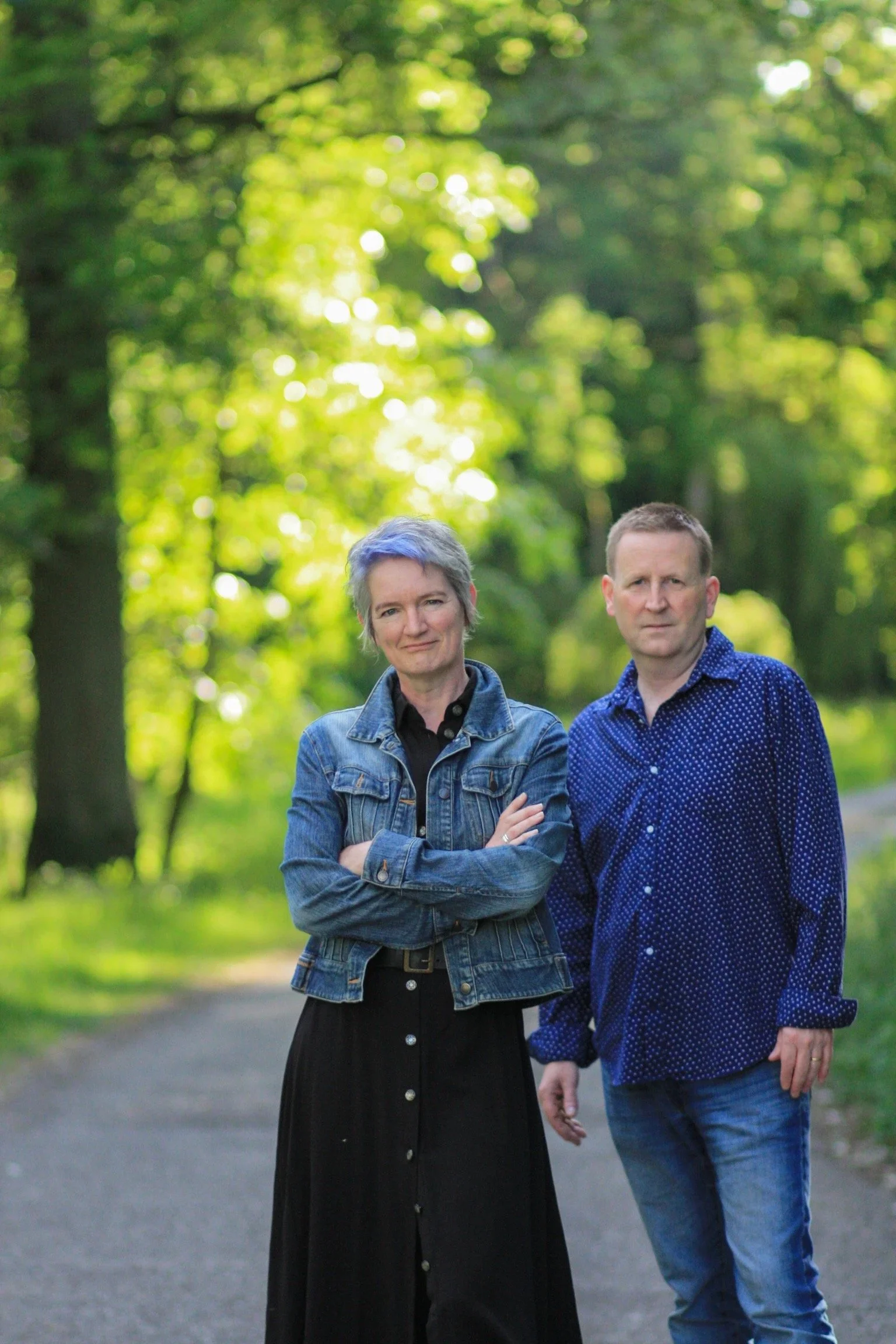 A woman and a man standing on a forest path with trees and sunlight in the background.