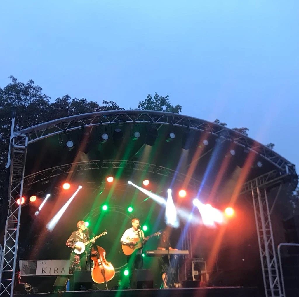 A live outdoor music concert at dusk, with a band performing on stage under colorful stage lighting, including two guitarists, a keyboardist, and a double bass, with trees visible in the background.