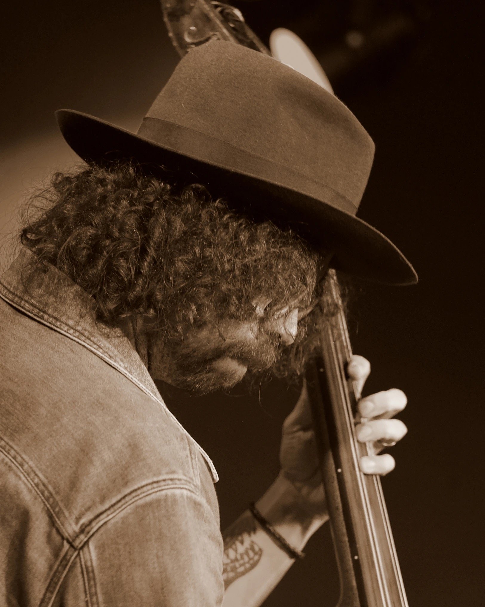 A man with curly hair and a beard, wearing a hat and a denim jacket, holding and kissing a guitar neck.