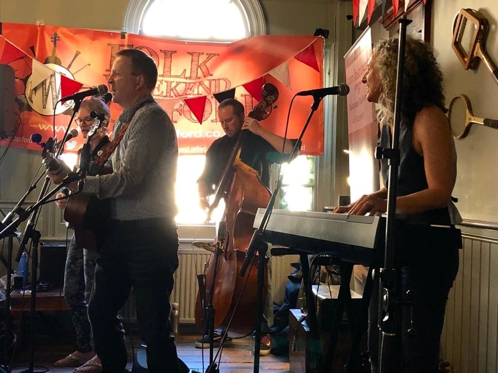 Musicians performing on stage at an event, with a banner reading 'Folk Weekend' in the background.