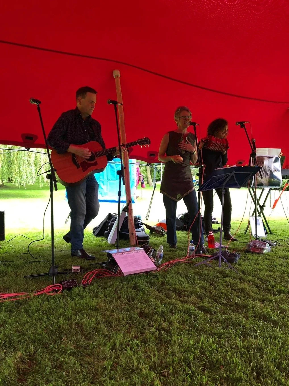 Three musicians performing under a red tent outdoors, with a man playing guitar, a woman singing, and another woman standing with a music stand. There are microphones, music sheets, and equipment on the grass.