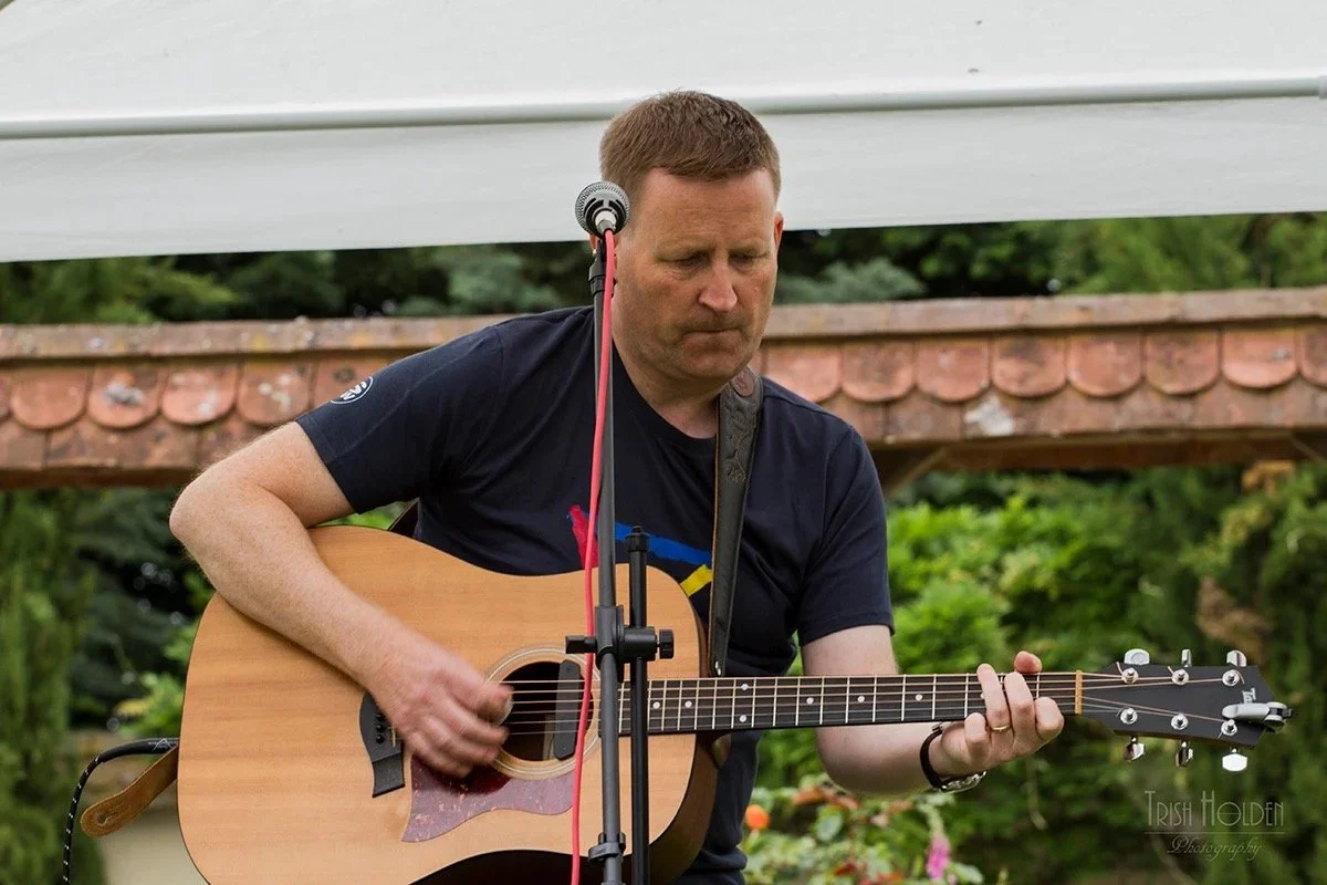 A man with short brown hair playing an acoustic guitar outdoors, with a microphone setup in front of him and a green landscape background.