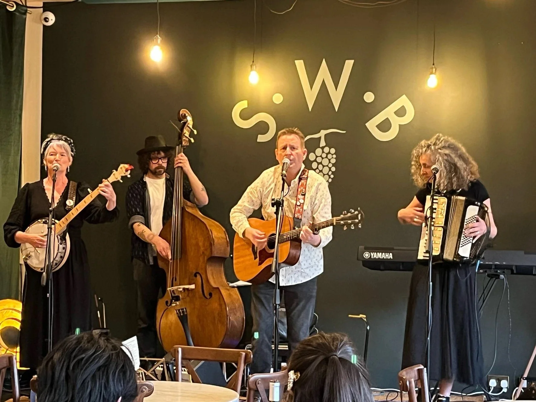 Four musicians performing on stage with a black wall backdrop. From left to right: a woman with short gray hair playing a banjo, a man with curly hair and glasses playing a double bass, a man with short hair singing and playing an acoustic guitar, an