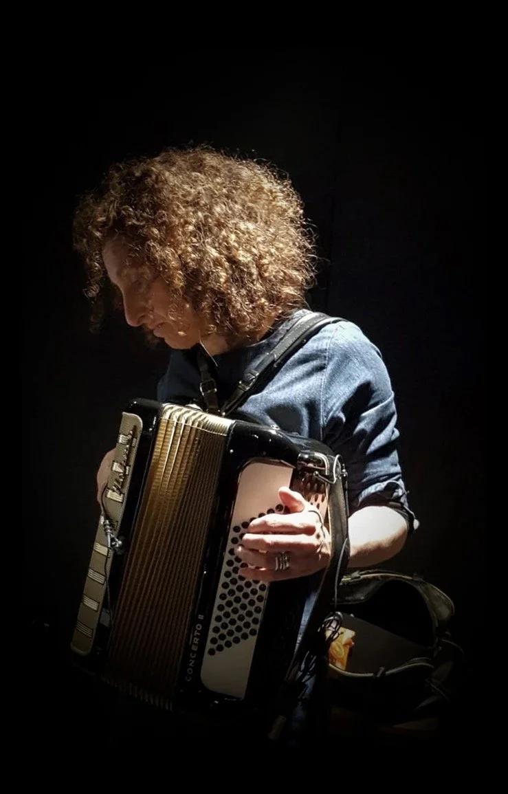 A person with curly hair wearing a denim shirt playing an accordion against a dark background.