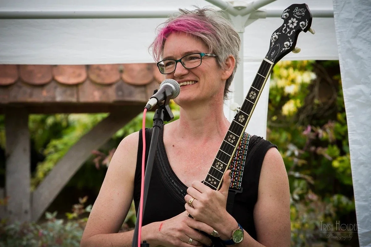 Woman with short, pink and gray hair, glasses, and a black sleeveless top, holding a guitar and smiling at a microphone during an outdoor event.