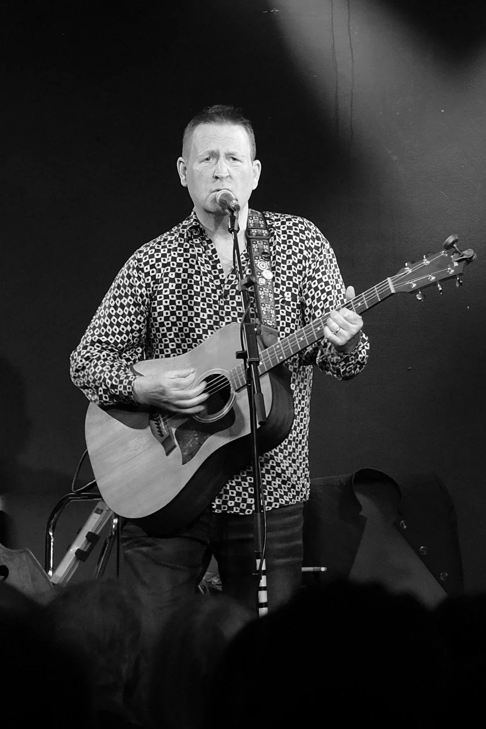 A man singing into a microphone while playing an acoustic guitar on stage, with a dark background.