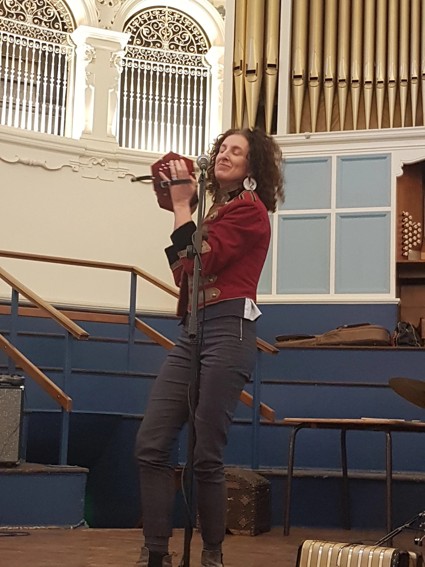 A woman with curly hair is singing or performing on stage, holding a microphone and a small red musical instrument, in a room with decorative architecture and an organ in the background.