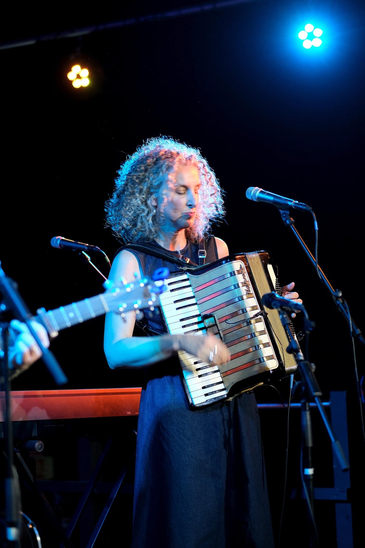 Female musician with curly gray hair in a black dress playing an accordion on stage with colorful stage lights.