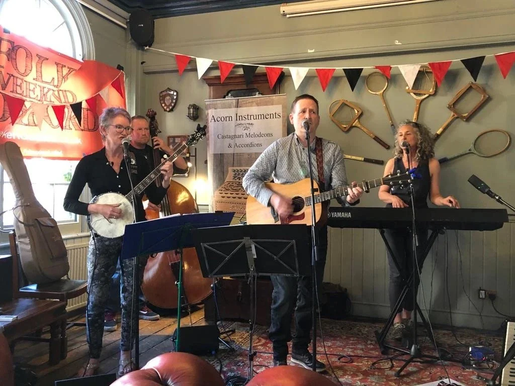 A four-piece band performing on stage at a local event, with a banjo, guitar, upright bass, and keyboard. Red, black, and white bunting hangs above, and a sign for 'Acorn Instruments' is visible in the background. The setting appears to be a cozy ind