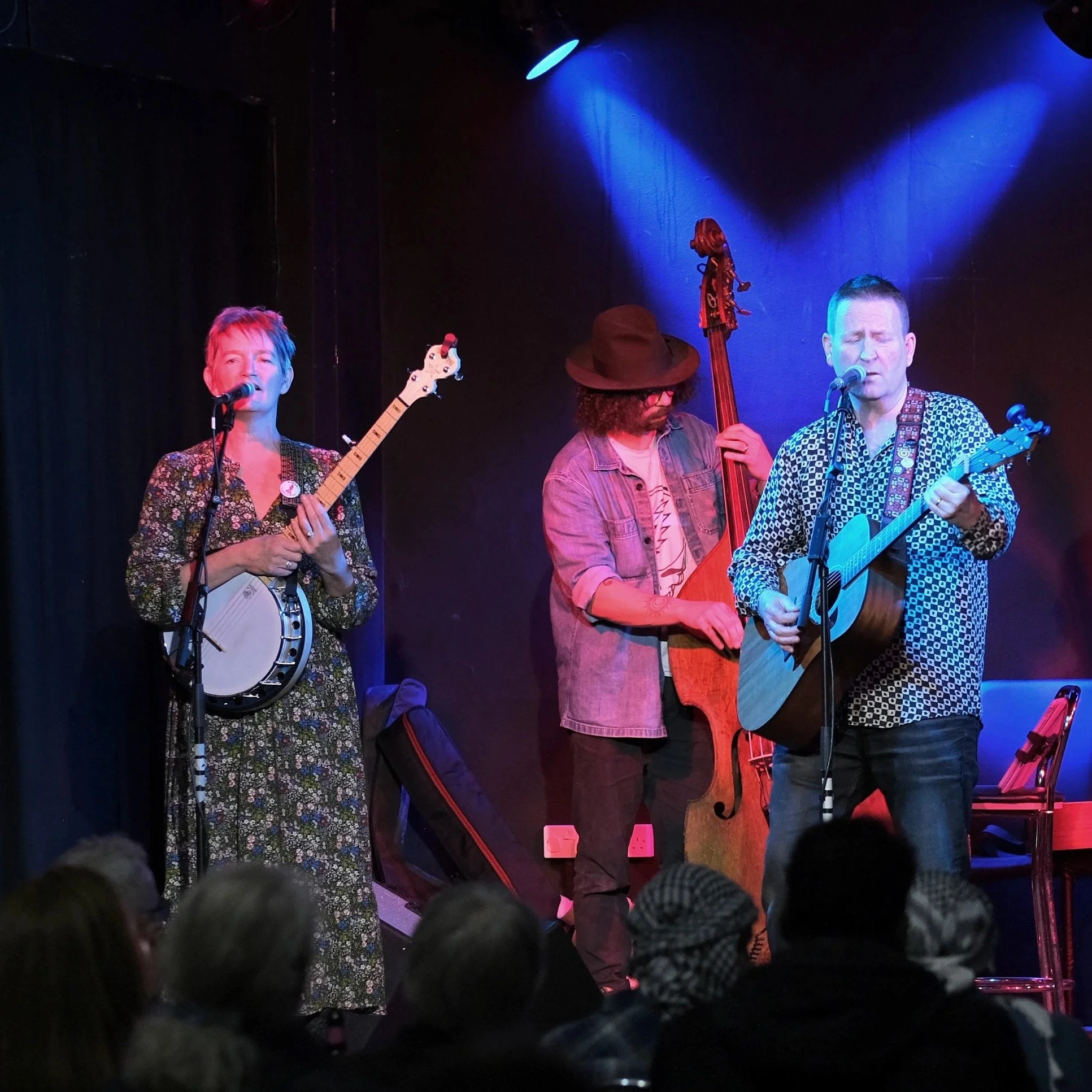 Three musicians performing on stage with dark background and blue stage lighting. The woman on the left is singing and playing a banjo, the man in the center is playing a double bass, and the man on the right is singing and playing an acoustic guitar