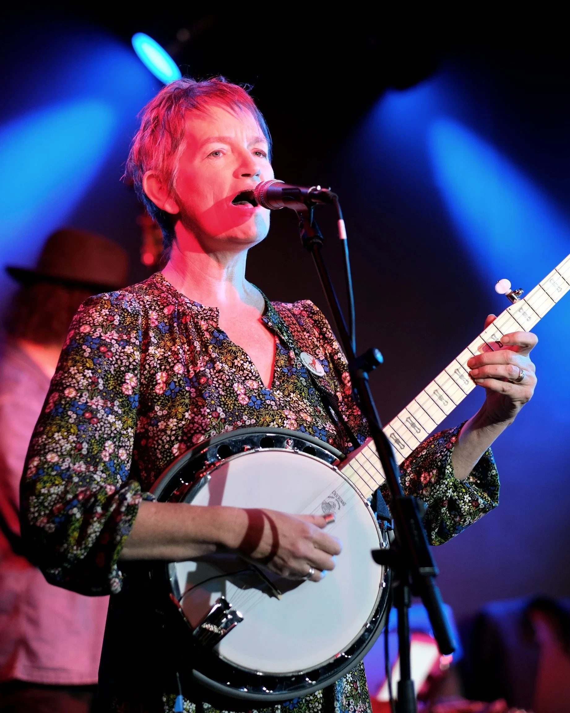 A woman with short hair sings into a microphone while playing a banjo on stage, with a musician and colorful stage lighting in the background.
