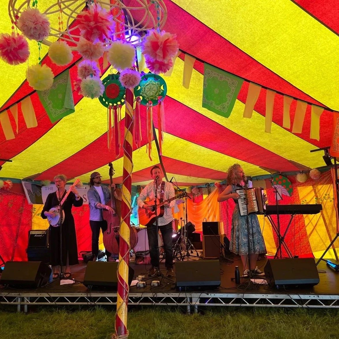 A live band performing on a decorated outdoor stage under a colorful striped tent, with musicians playing instruments including a banjo, double bass, guitar, and accordion.