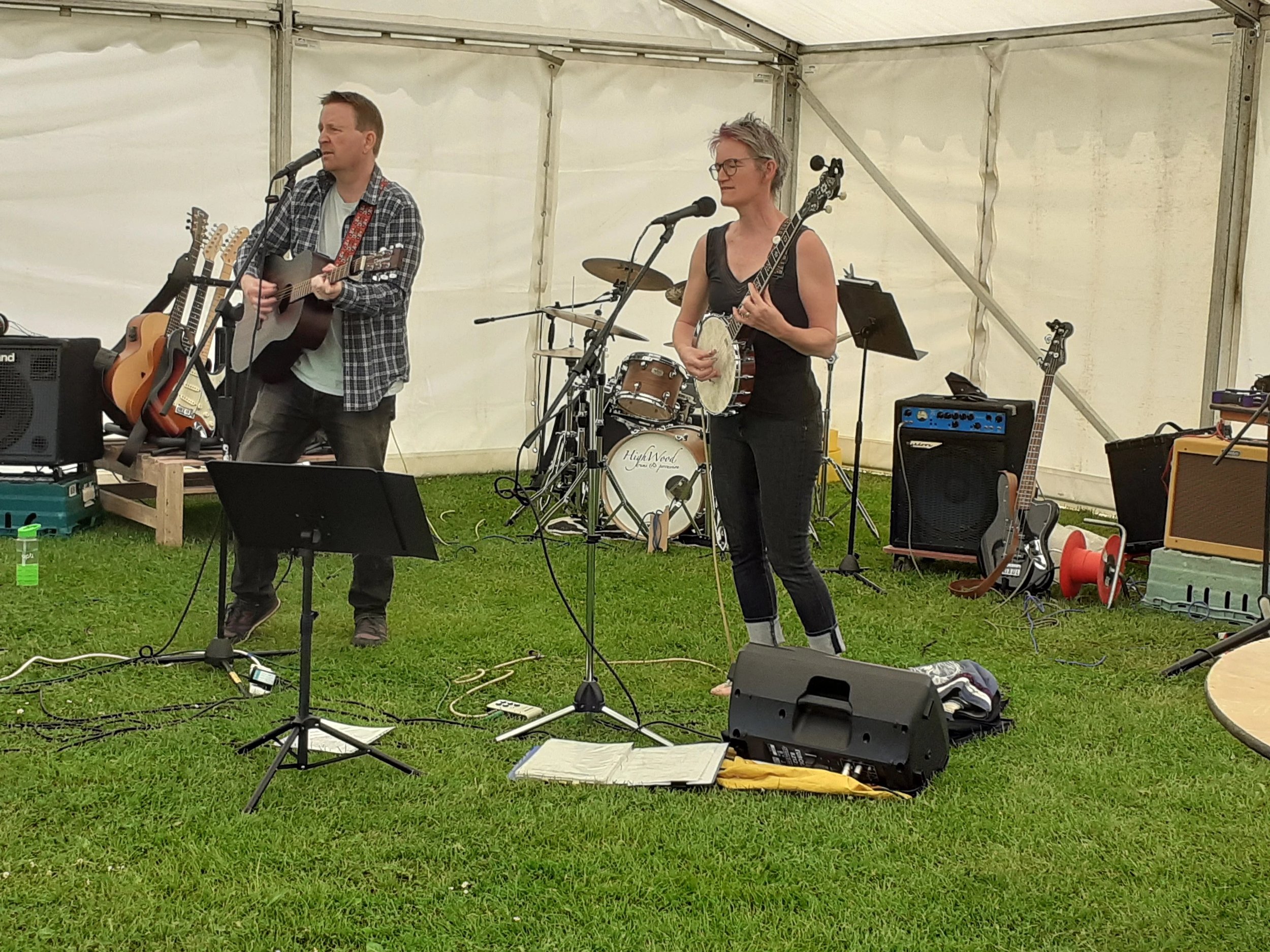 Two musicians, a man and a woman, perform on stage inside a white tent. The man plays an acoustic guitar and sings into a microphone, while the woman plays a mandolin and also sings into a microphone. Various musical equipment, including guitars, amp