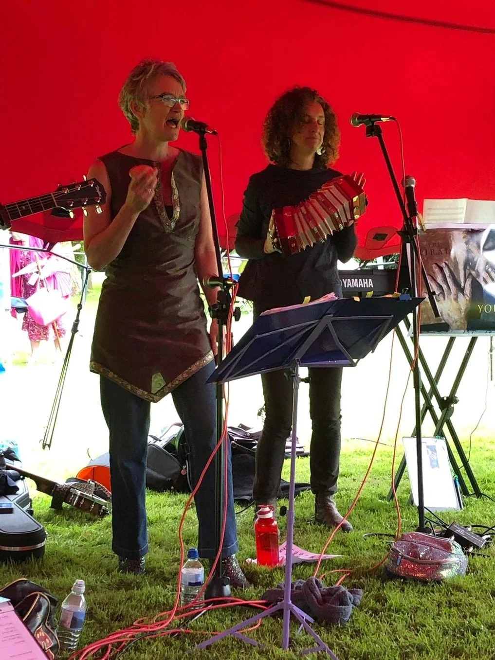 Two women are performing music under a red canopy. One woman is singing into a microphone, holding a guitar, and the other is playing a cajón. They are surrounded by music stands, a keyboard, and various musical equipment.