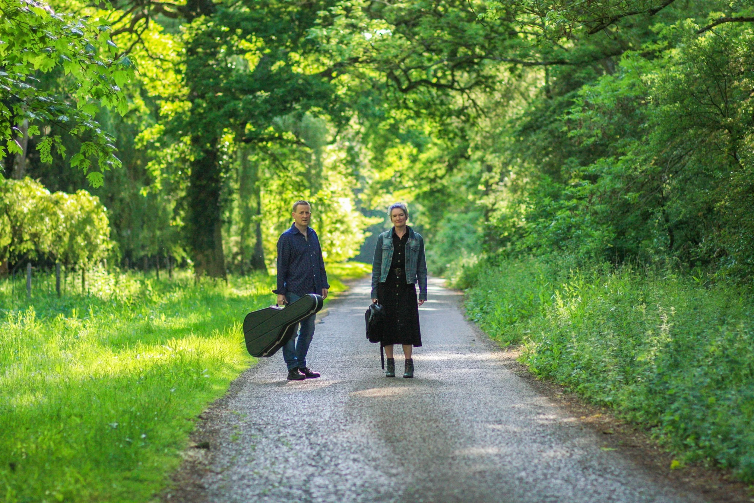 A man and woman walking on a dirt path surrounded by green trees and grass, with the man carrying a guitar case.