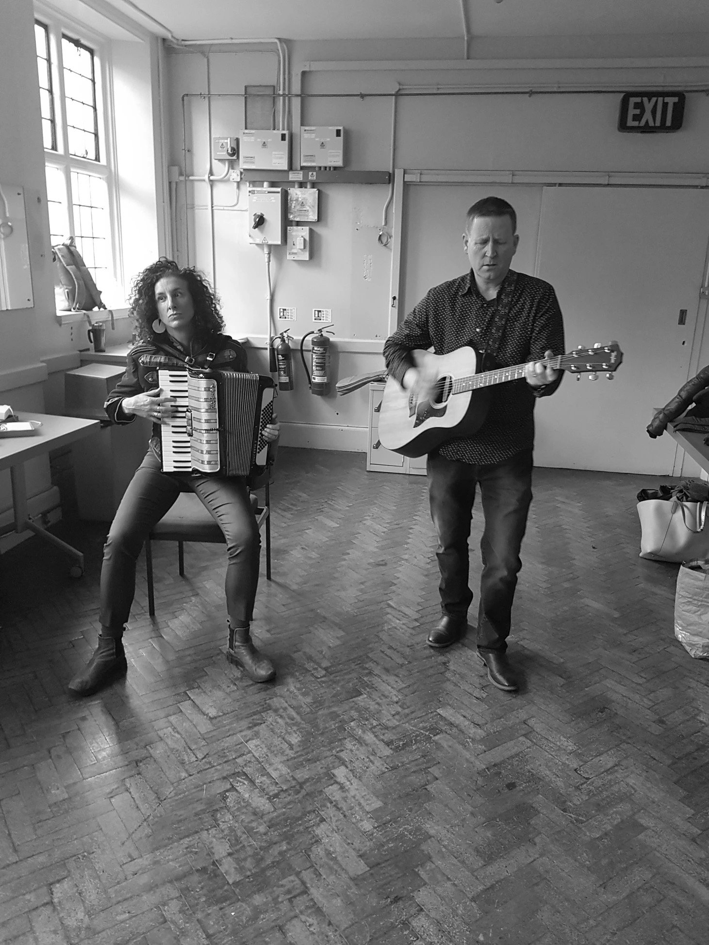 A woman playing an accordion and a man playing an acoustic guitar, performing indoors in a room with large windows and an EXIT sign.