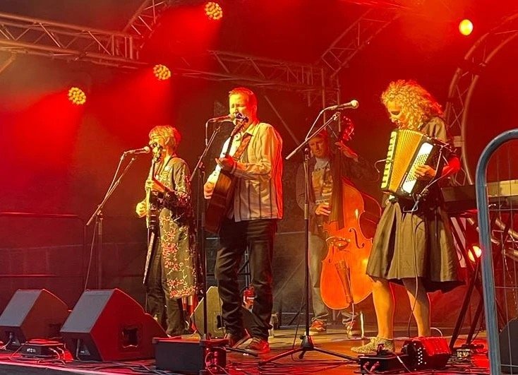 A band of five musicians performing on stage with red lighting, including a female accordion player, a male guitarist singing and playing, a male upright bass player, and two female singers with microphones.