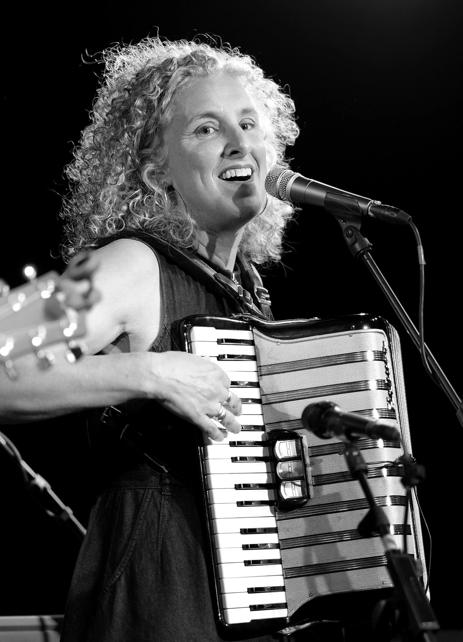 A woman with curly hair singing into a microphone while playing an accordion.
